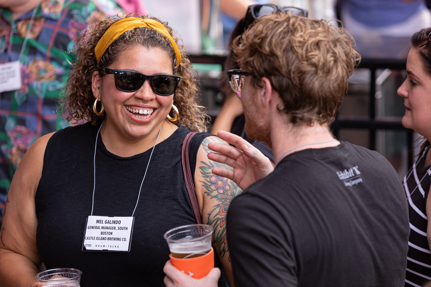 A laughing woman in sunglasses, a yellow headband and a black sleeveless top with a name badge talks with a man in a black Exhibit A Brewing Company t-shirt, cups of beer in hand.