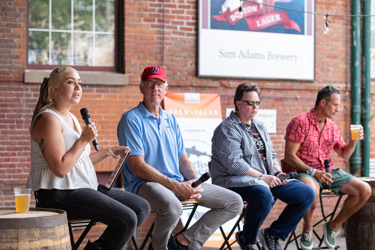 A woman with blonde braids in a white sleeveless top speaks into a microphone beside a man in a red W cap and light blue polo, a person in sunglasses and a gingham shirt, and a man in a red patterned shirt holding a beer, in front of a brick wall and a Sam Adams sign, with a glass of beer on a barrel at the left.