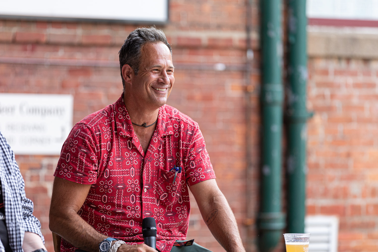 A smiling man in a red patterned short-sleeve shirt with a microphone in his lap and a cup of beer beside him, in front of a brick wall with green drainpipes.