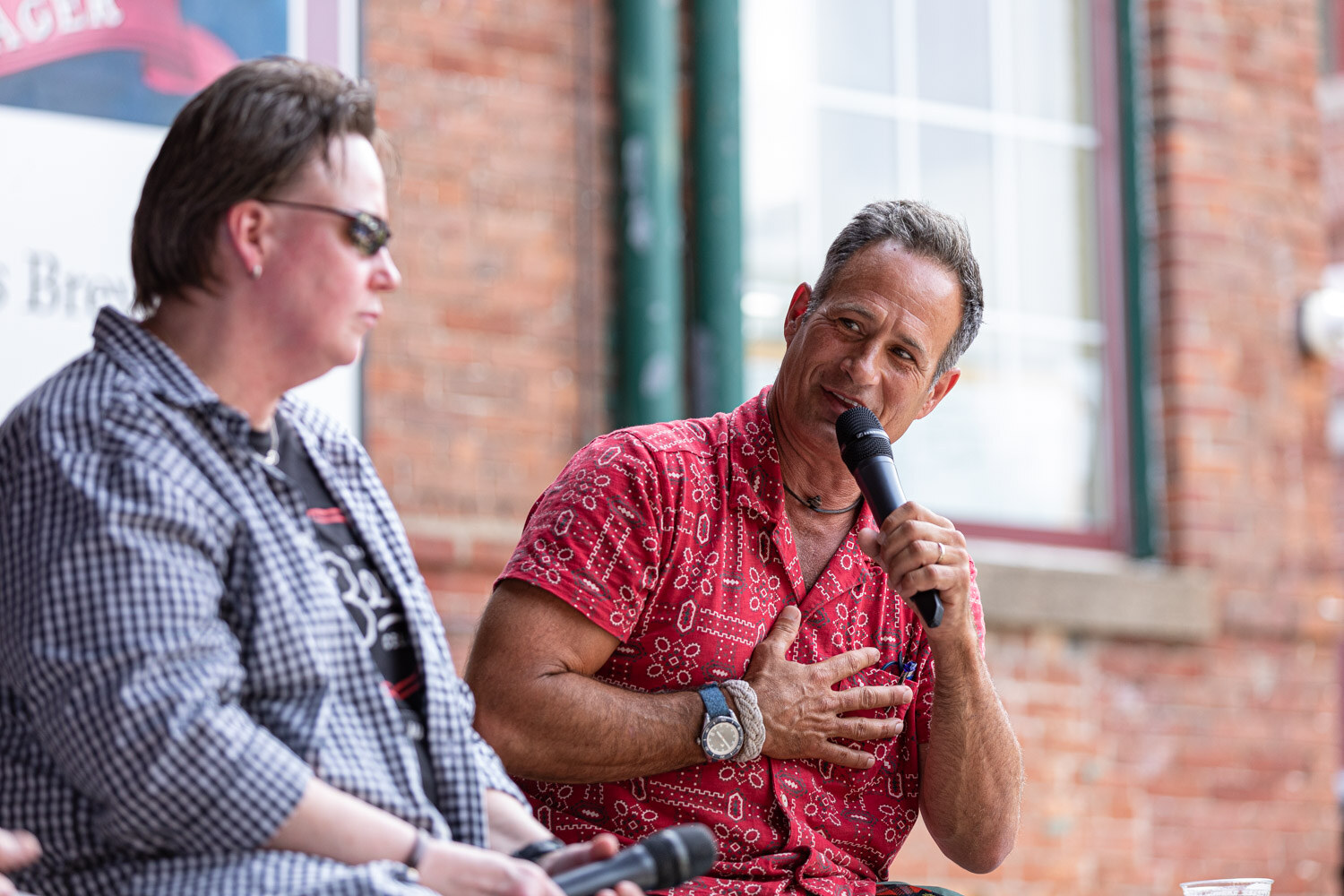A man in a red patterned shirt speaks into a microphone with a hand on his chest while a person in sunglasses and a gingham shirt sits in the foreground with microphones in their lap.