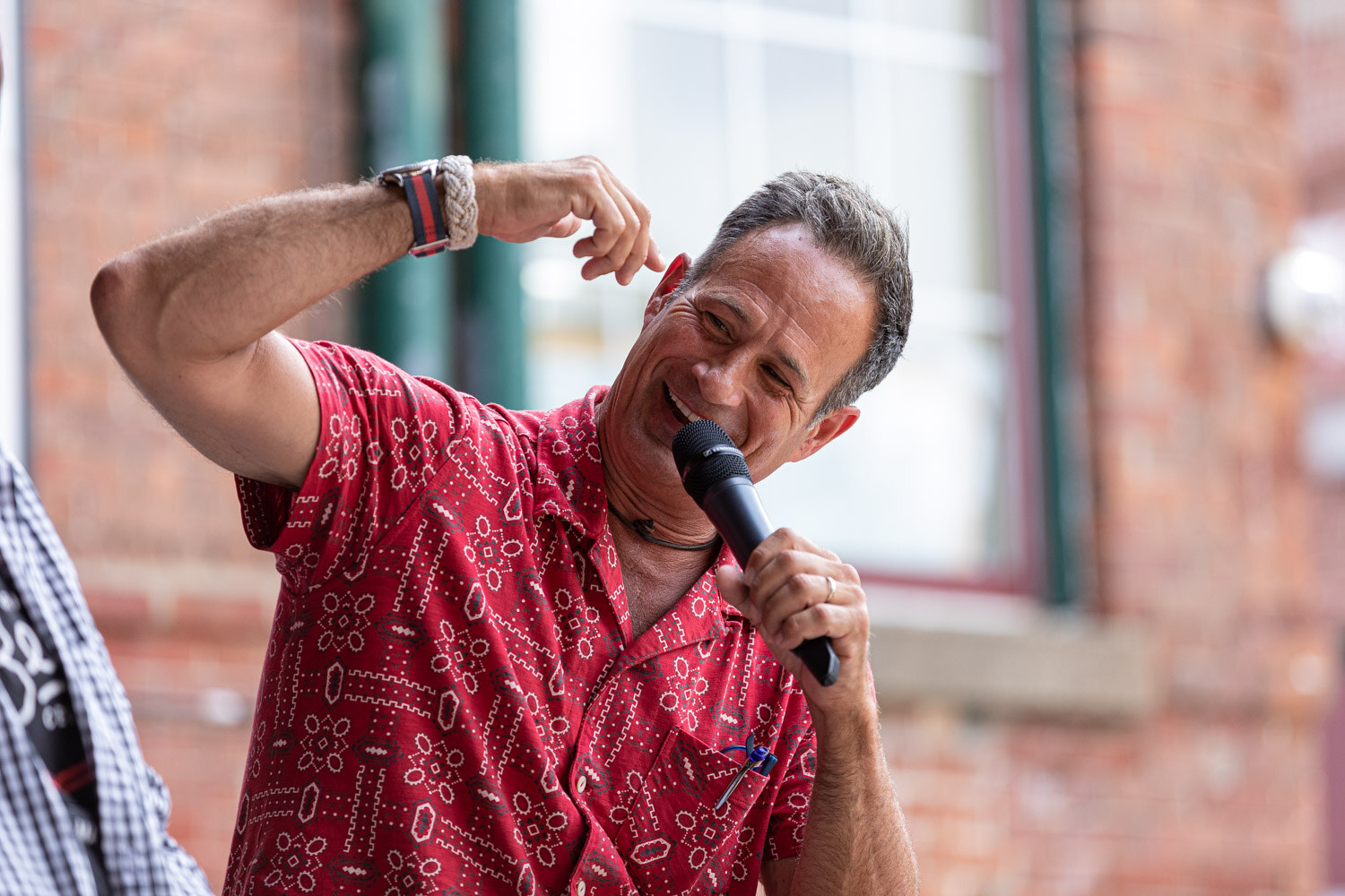 A man in a red patterned shirt laughs with a microphone at his mouth and one hand raised near his ear, in front of a blurred brick wall and window.