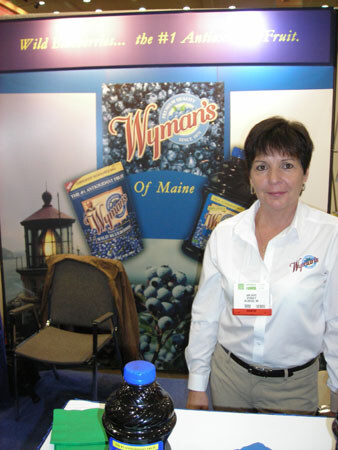 A woman in a white Wyman's shirt stands beside a Wyman's of Maine wild blueberry backdrop, with a bottle of dark juice on the table in front.