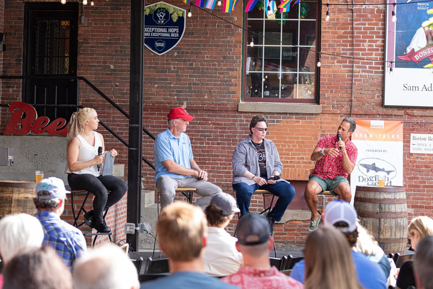 Four people on stools in front of a red brick wall with a red Beer sign and a Brew Talks banner presented by Dogfish Head, with a woman in a white top at left and a man in a red patterned shirt speaking at right, and audience heads in the foreground.