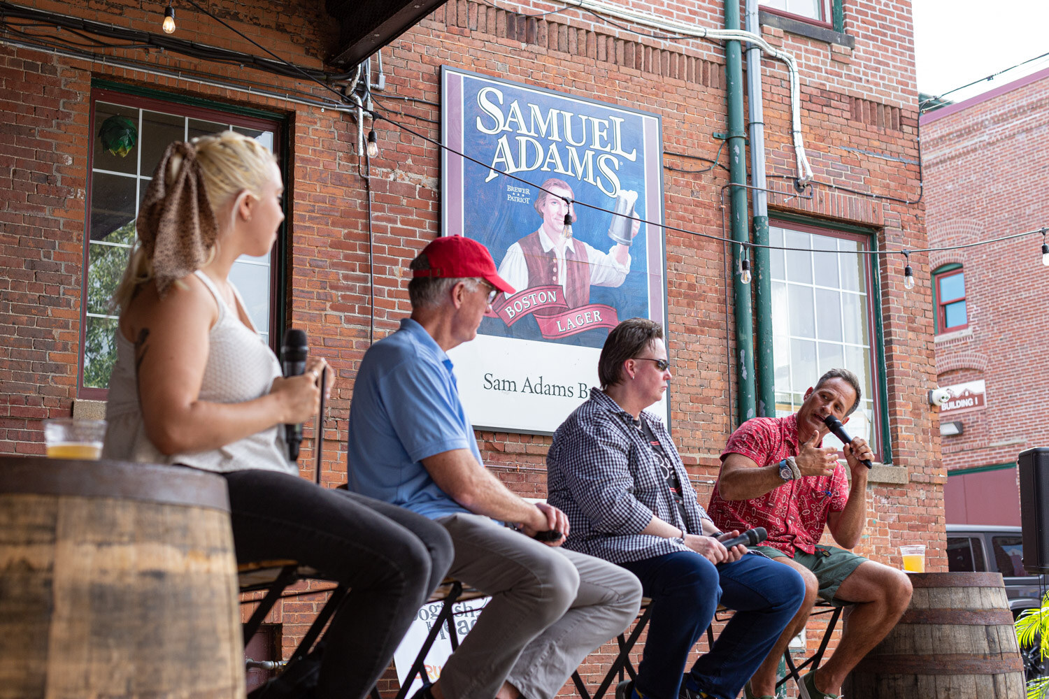 Four people on stools in front of a brick wall under a Samuel Adams Boston Lager sign, with a woman in a white top and hair scarf holding a microphone in the foreground at left and a man in a red patterned shirt speaking at right, with a barrel at each side.
