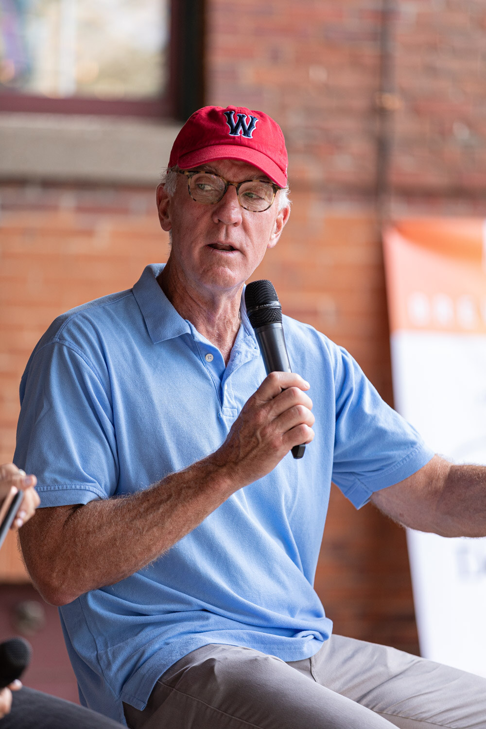 Portrait-format shot of a man in a red cap with a W logo, round glasses and a light blue polo speaking into a microphone in front of a brick wall and an orange banner.