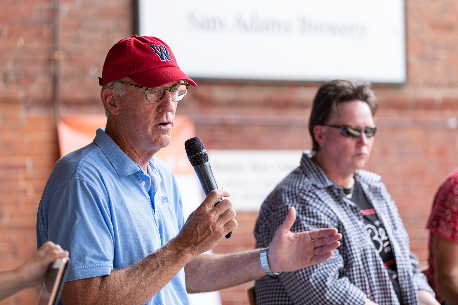 A man in a red W cap, glasses and a light blue Harpoon polo gestures with one hand while holding a microphone, with a person in sunglasses and a gingham shirt behind him.