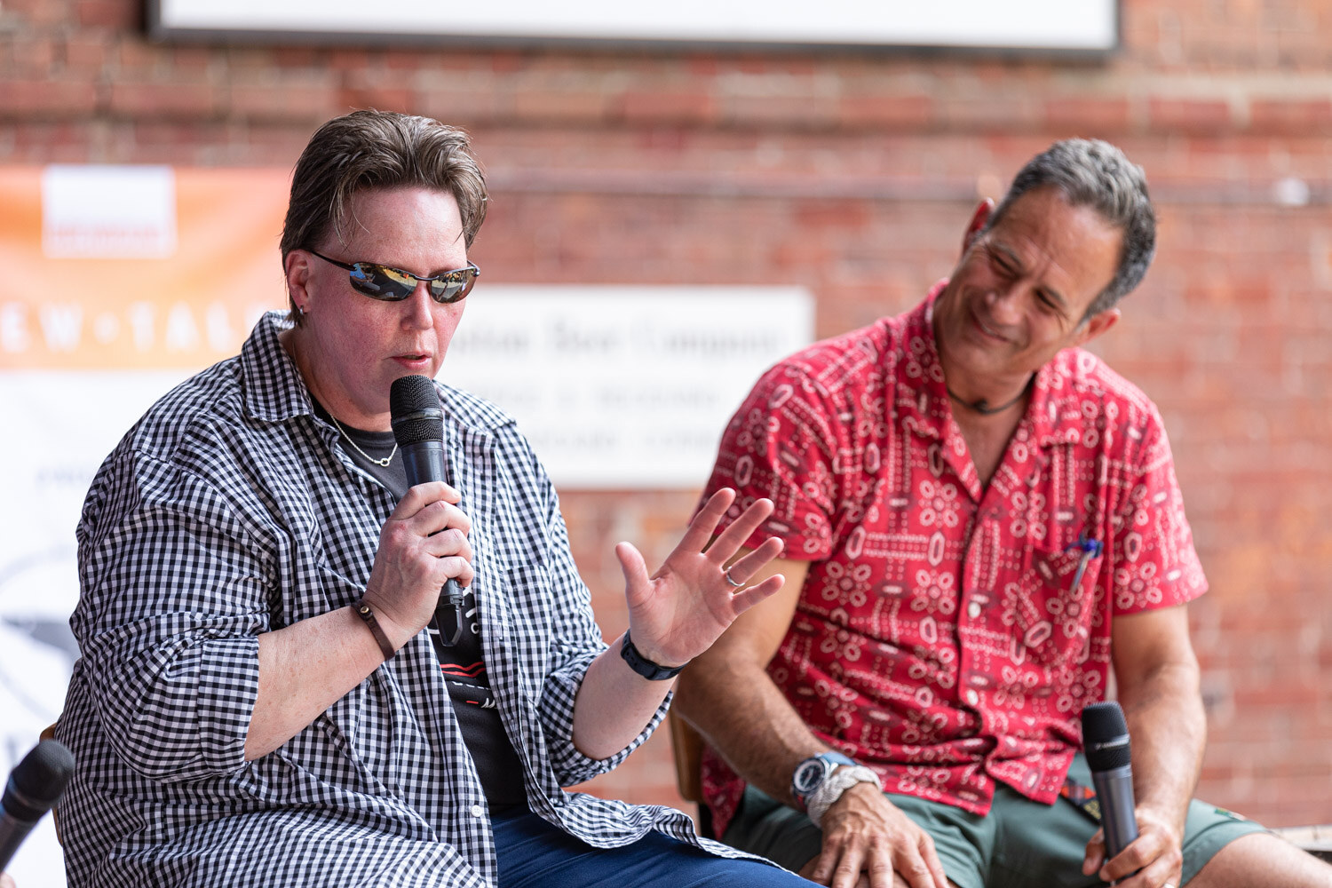 A person in sunglasses and a black and white gingham shirt over a dark graphic t-shirt speaks into a microphone with an open hand while a man in a red patterned shirt looks down and smiles.