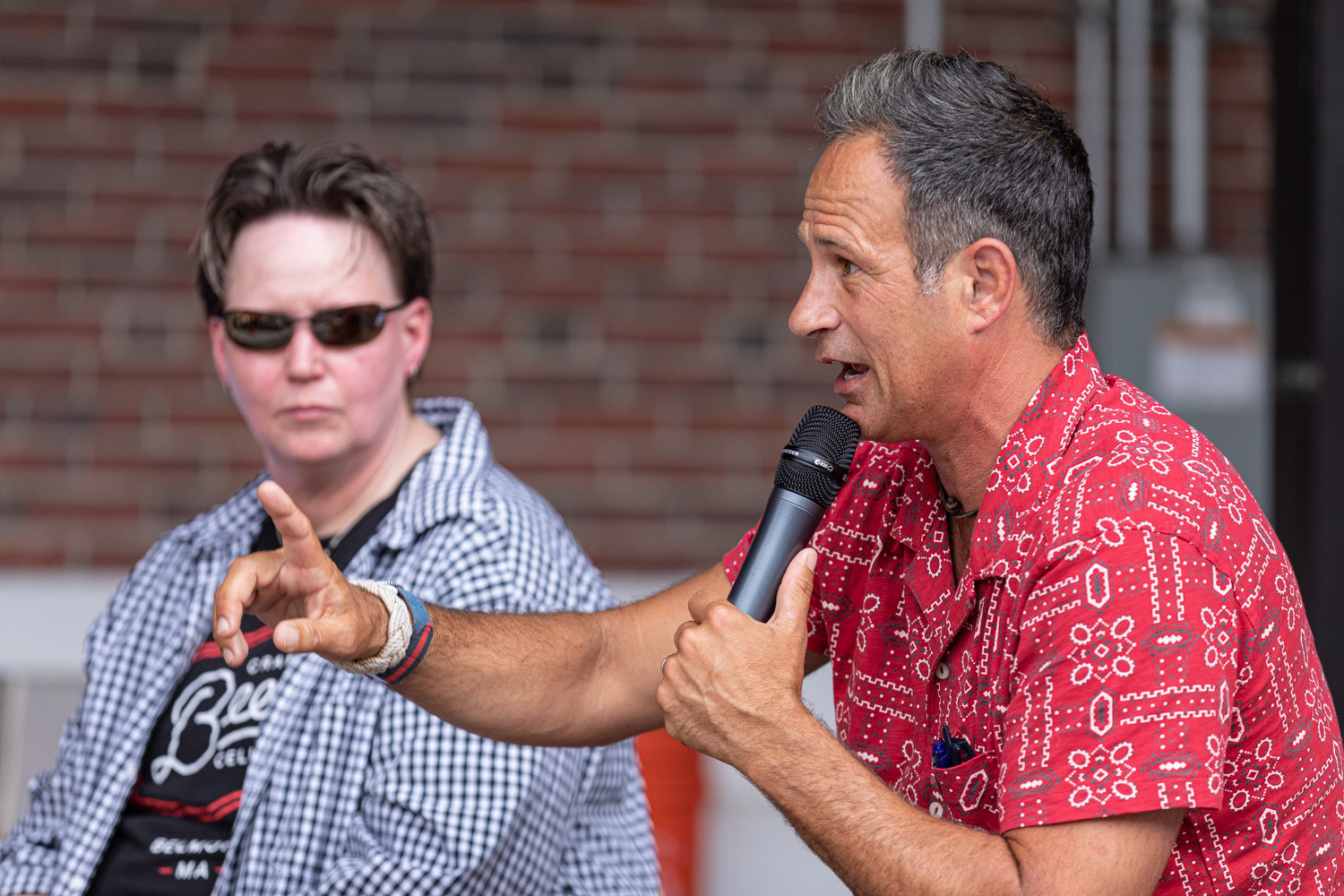 A man in a red patterned shirt points with one hand and holds a microphone in the other while a person in sunglasses and a gingham shirt looks toward him.