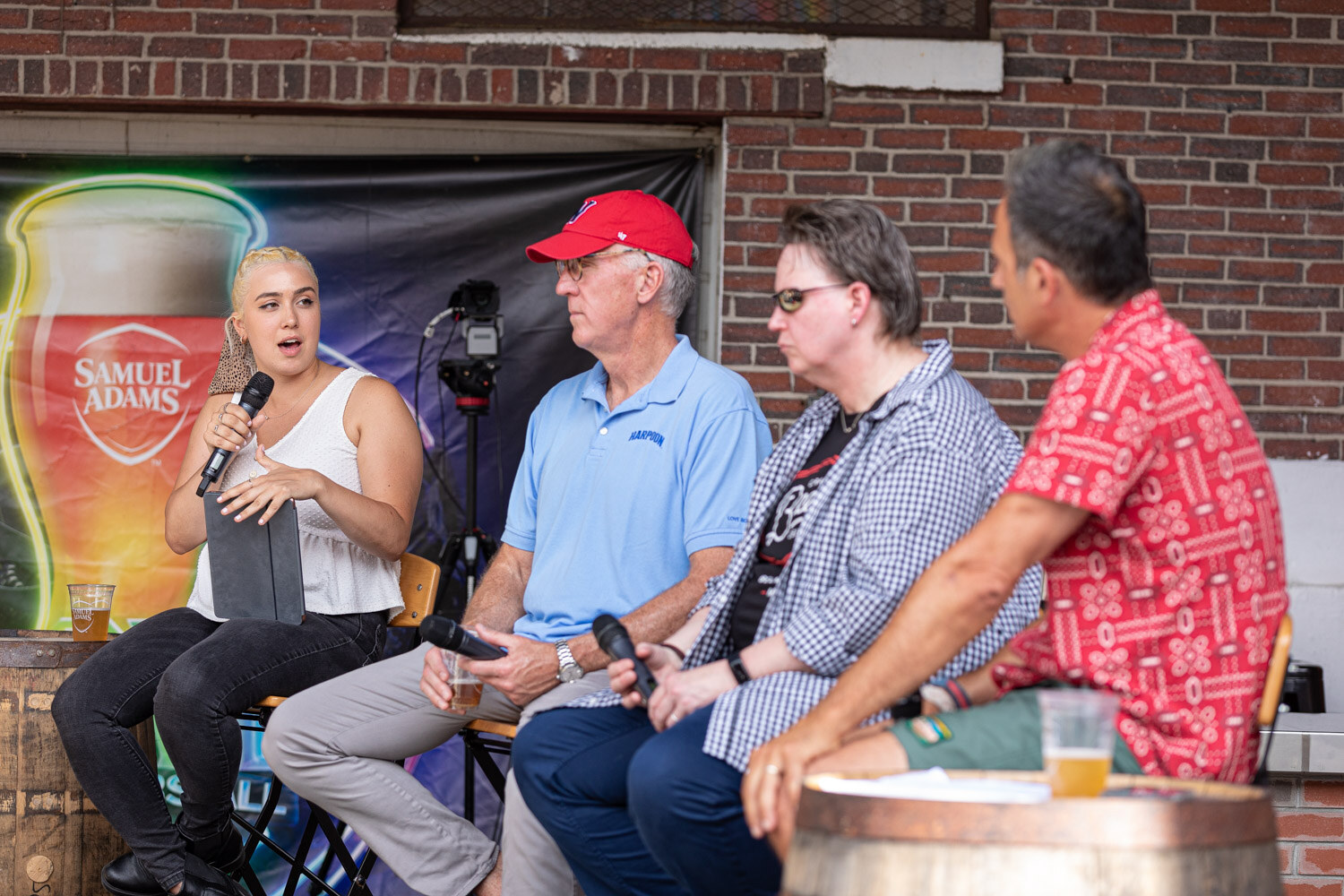 A woman in a white top holding a tablet and microphone speaks to a man in a red W cap and blue Harpoon polo, a person in a gingham shirt and a man in a red patterned shirt, with a video camera on a tripod and a neon Samuel Adams glass banner behind them.