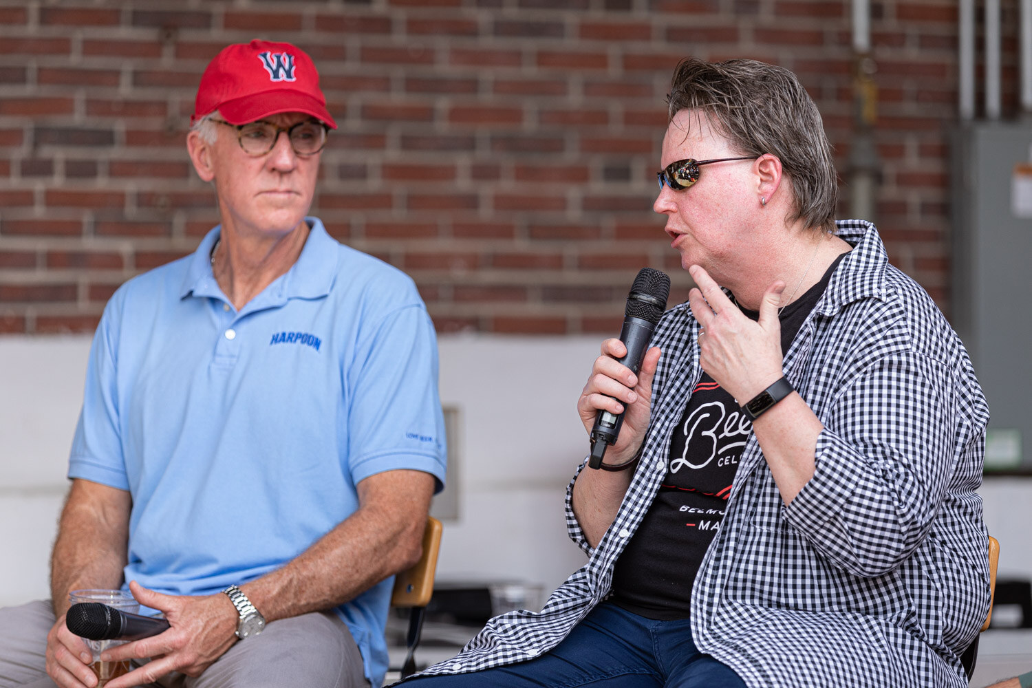 A person in sunglasses and a gingham shirt over a black graphic t-shirt speaks into a microphone while a man in a red W cap and a light blue Harpoon polo listens beside her.