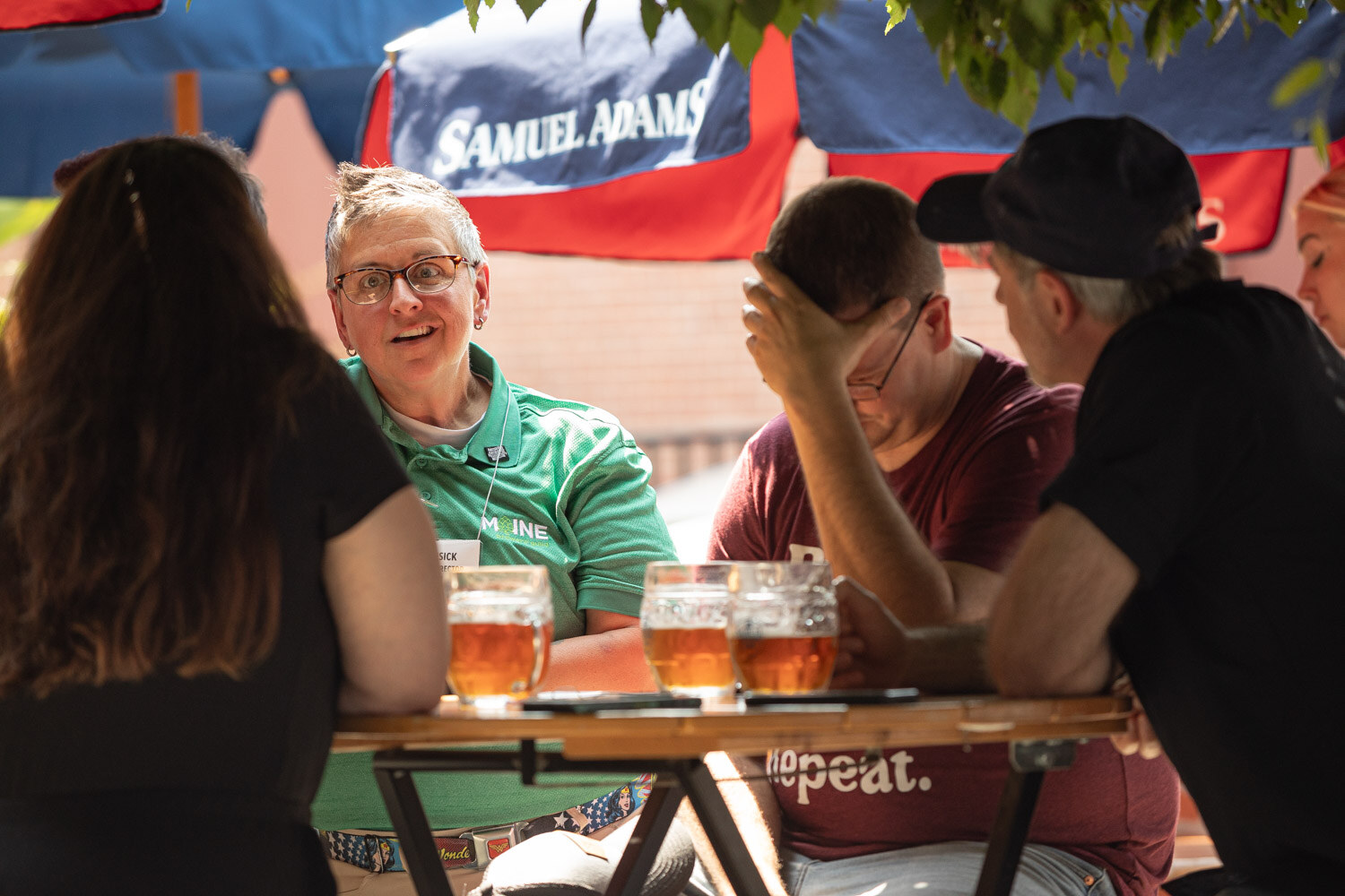 Four people talk around a wooden table with several glasses of amber beer under green leaves and a Samuel Adams umbrella.