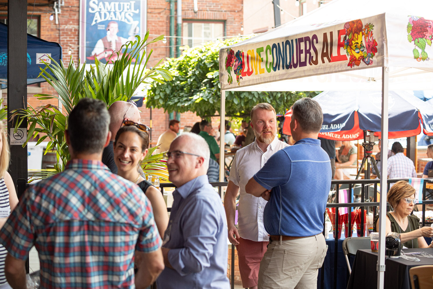Guests talk beneath a banner reading Love Conquers Ale with plants and a Samuel Adams sign in the background.