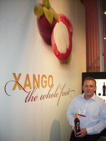 A man in a white shirt and badge holds a dark Xango bottle beside a wall graphic of mangosteen fruit and the text 'Xango the whole fruit.'