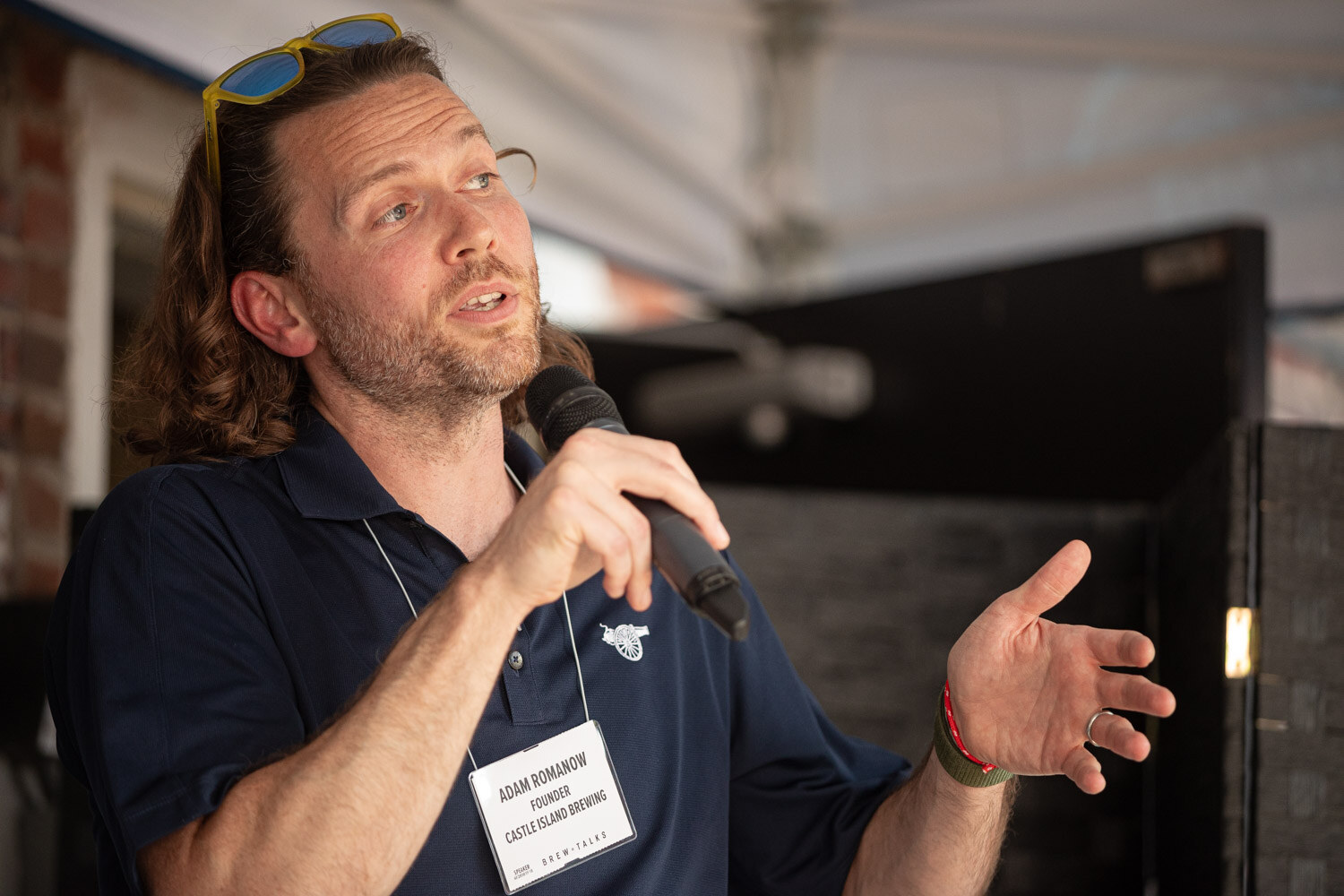 A man with long hair and sunglasses on his head speaks into a microphone and gestures under a white tent.