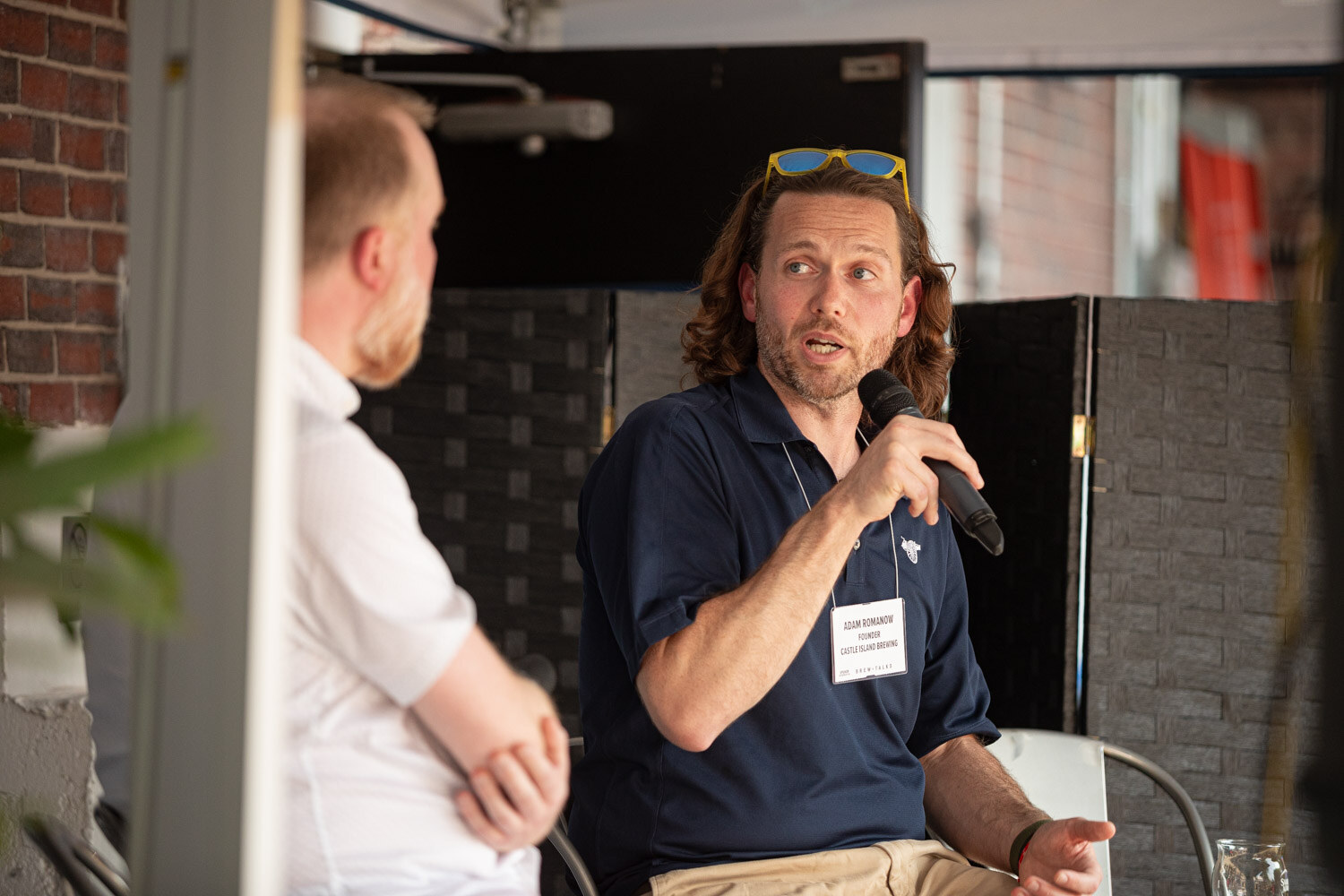 A man in a navy polo seated on a stool speaks into a microphone while a man in a white shirt sits next to him.