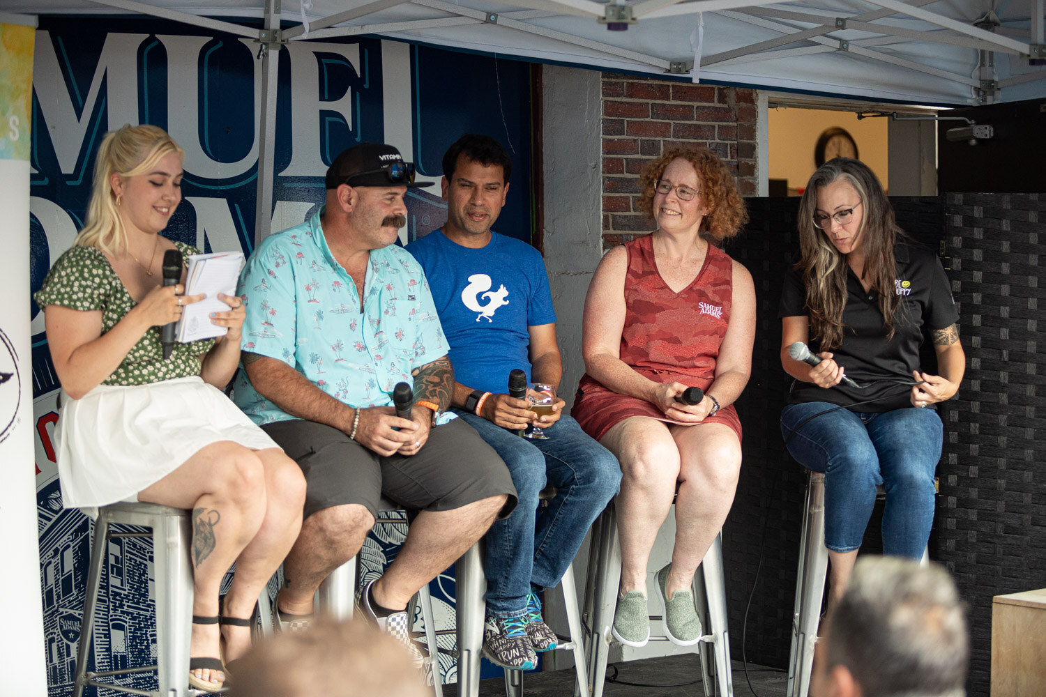Five panelists on tall stools hold microphones on a stage in front of a Samuel Adams backdrop.