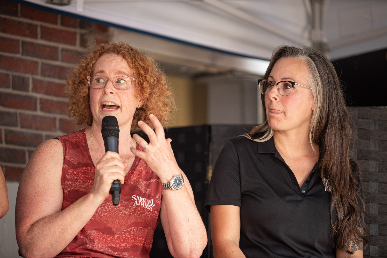 A woman with curly red hair in a Samuel Adams shirt speaks into a microphone while a woman in a black polo looks at her.