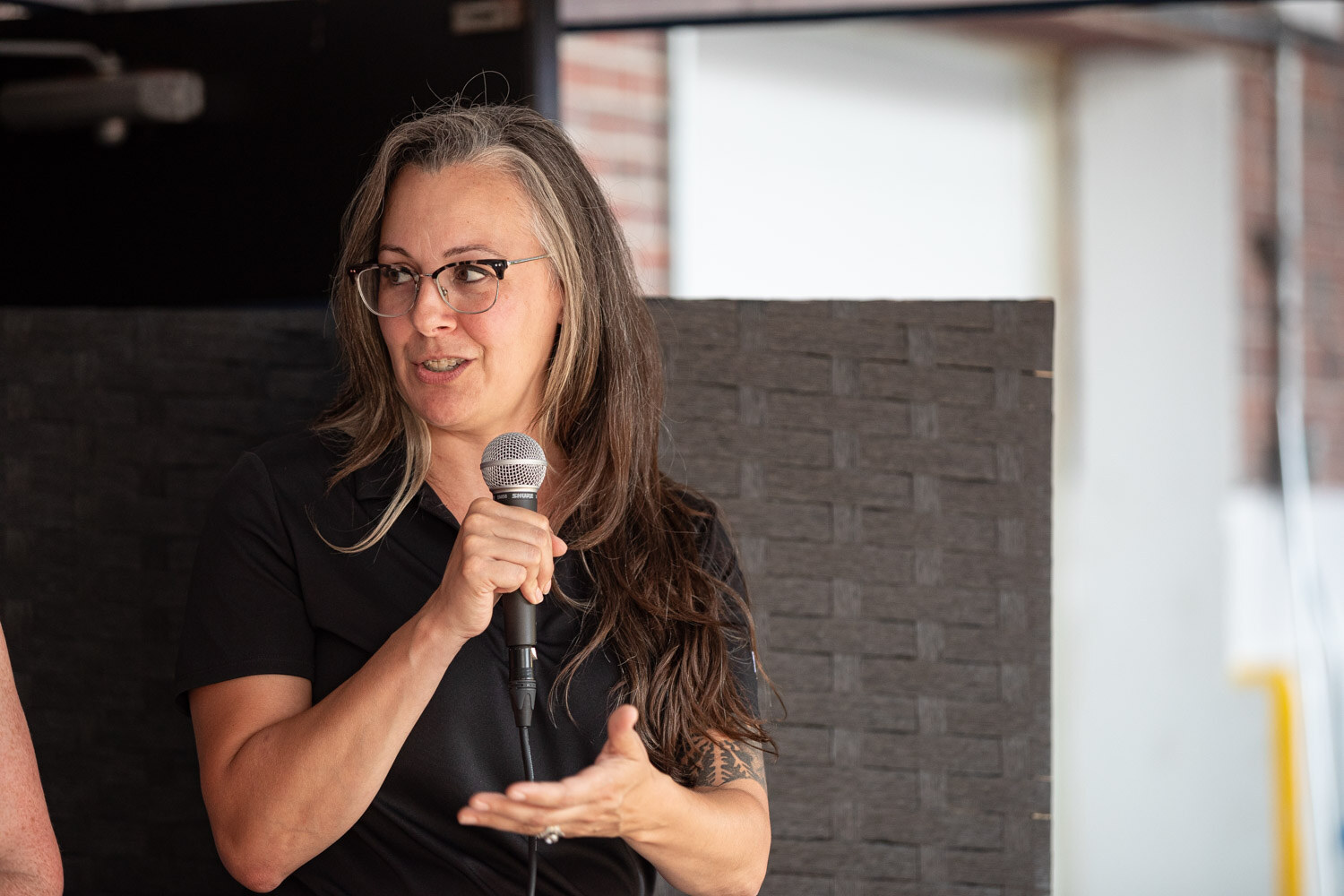 A woman with long gray hair and glasses speaks into a microphone and gestures with one hand.