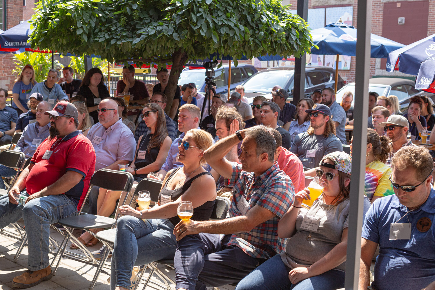 Seated audience under trees and a blue Samuel Adams umbrella, many holding beers.