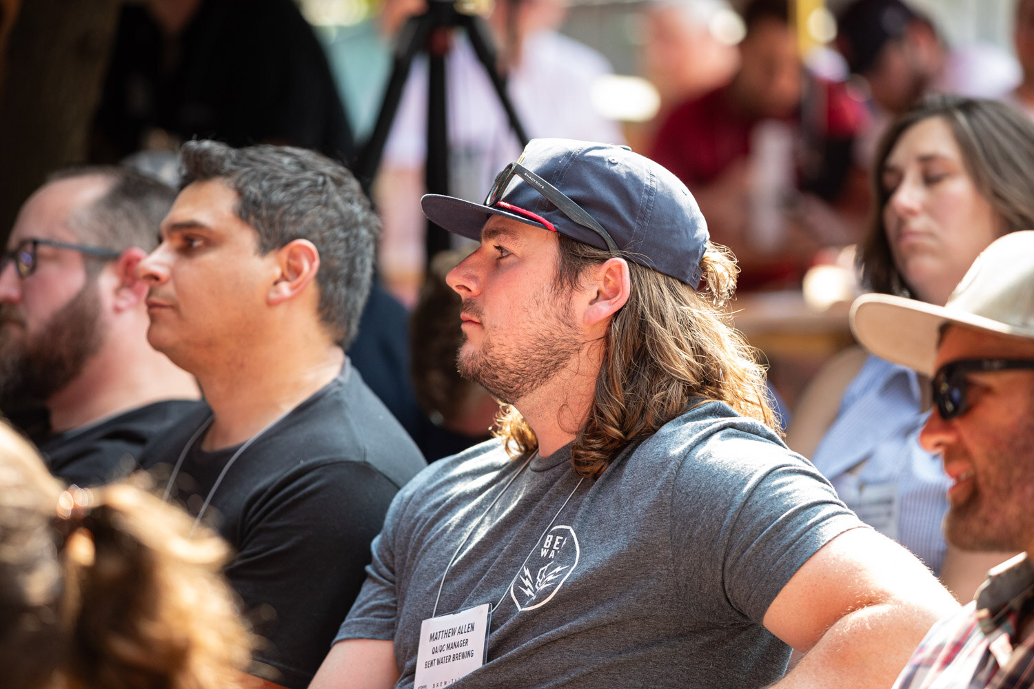 Profile view of a long-haired man in a baseball cap and gray shirt listening to a talk in a crowd.