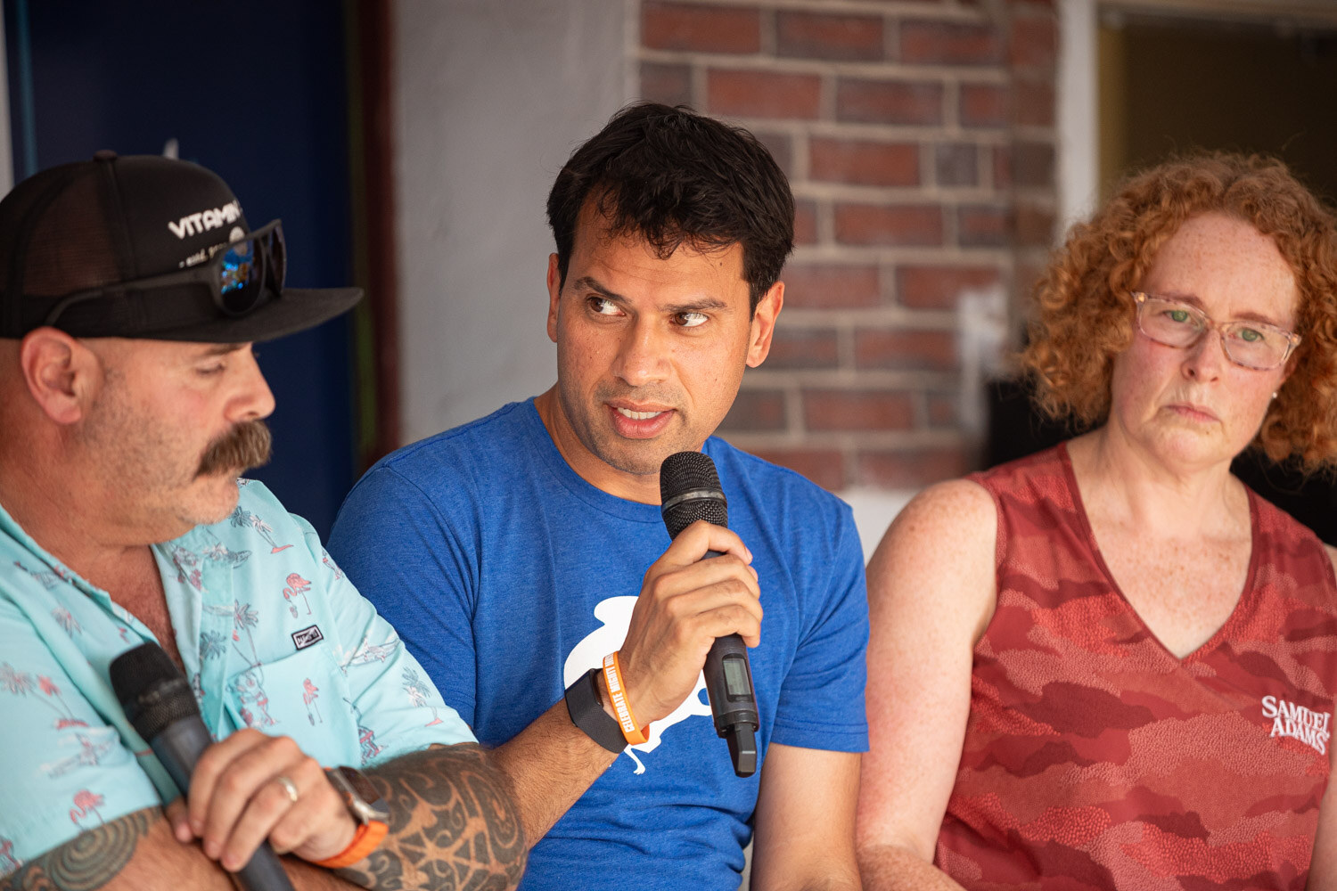 A man in a blue shirt speaks into a microphone next to a man in a patterned shirt and a woman with red curly hair.