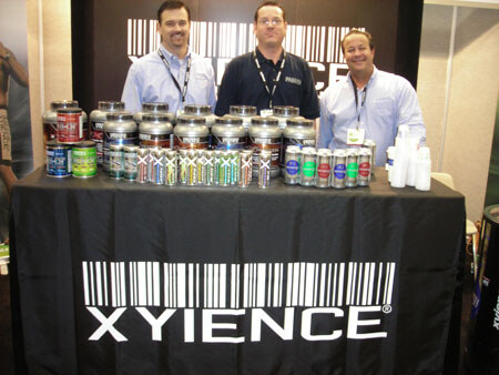 Three men stand behind a black table with a Xyience barcode logo, displaying tubs of supplements and rows of small cans and bottles.