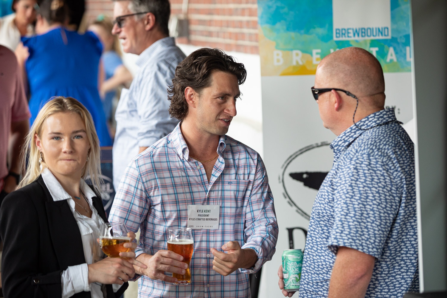 A man in a plaid shirt holds a beer and talks with a bald man in sunglasses and a woman beside a Brewbound banner.