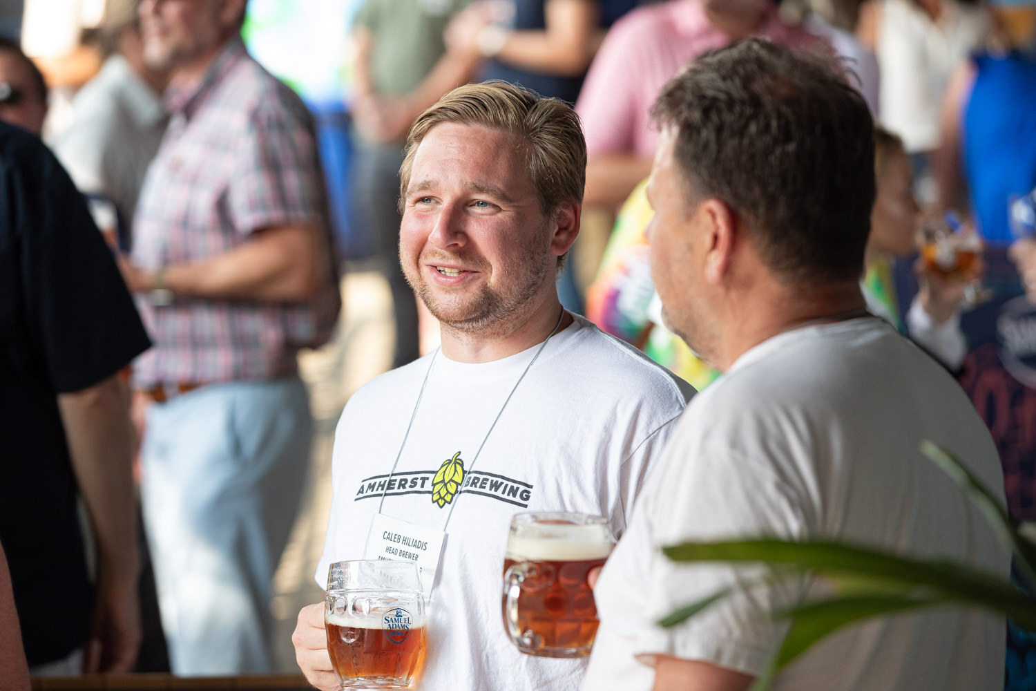 A smiling man in a white Amherst Brewing shirt holds a glass beer mug in a crowd of guests.