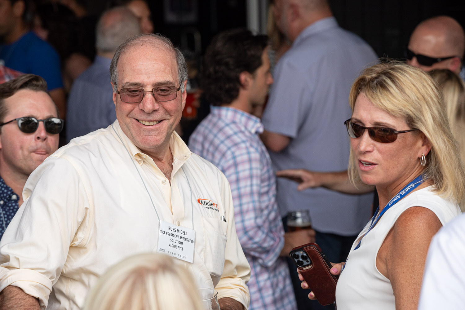 A smiling older man in sunglasses and a lanyard talks with a woman in a sleeveless top in a crowd of guests.