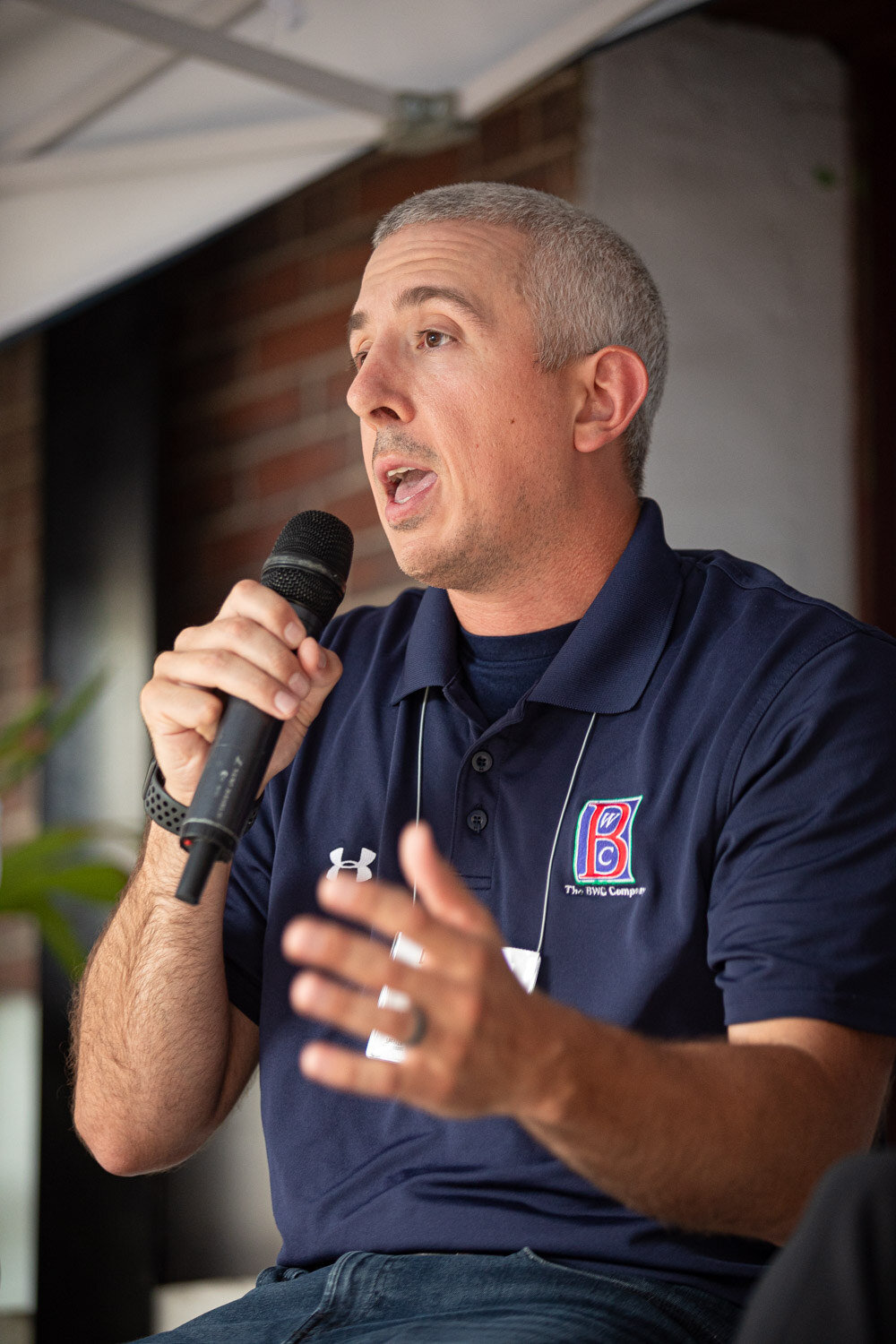 A man in a navy polo with a B logo speaks into a microphone and gestures with his hand.