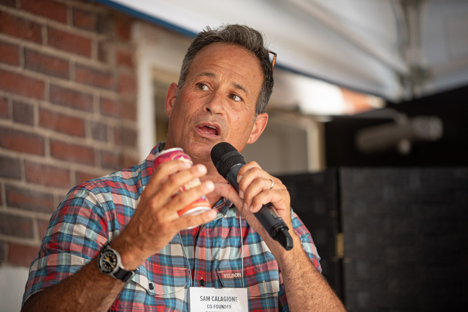 A man in a plaid shirt holding a red can speaks into a microphone.