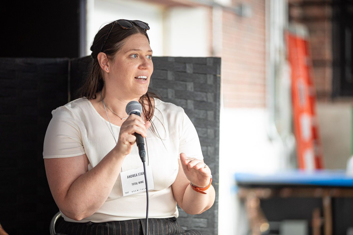A woman in a white top with a lanyard speaks into a microphone and gestures with her hand.