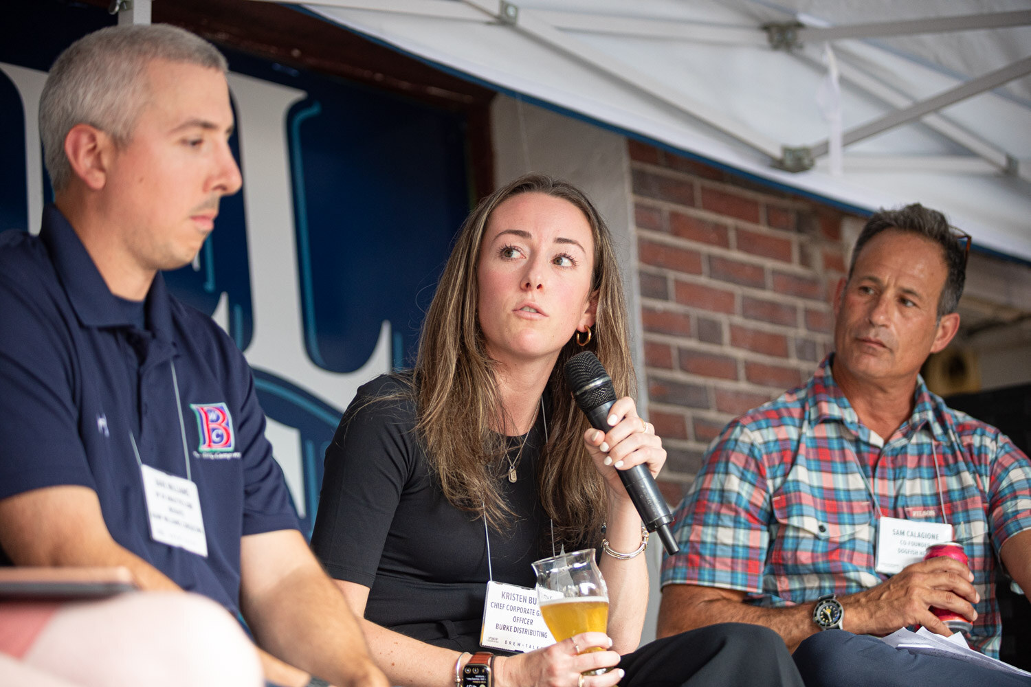 Three panelists on stage under a white tent, a man in a navy polo, a woman in black holding a beer, and a man in a plaid shirt with a can.