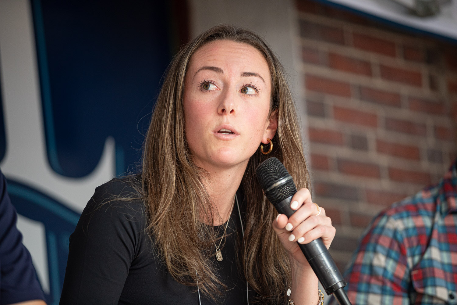 A woman with brown hair holds a microphone and looks to the side with a Samuel Adams backdrop behind her.