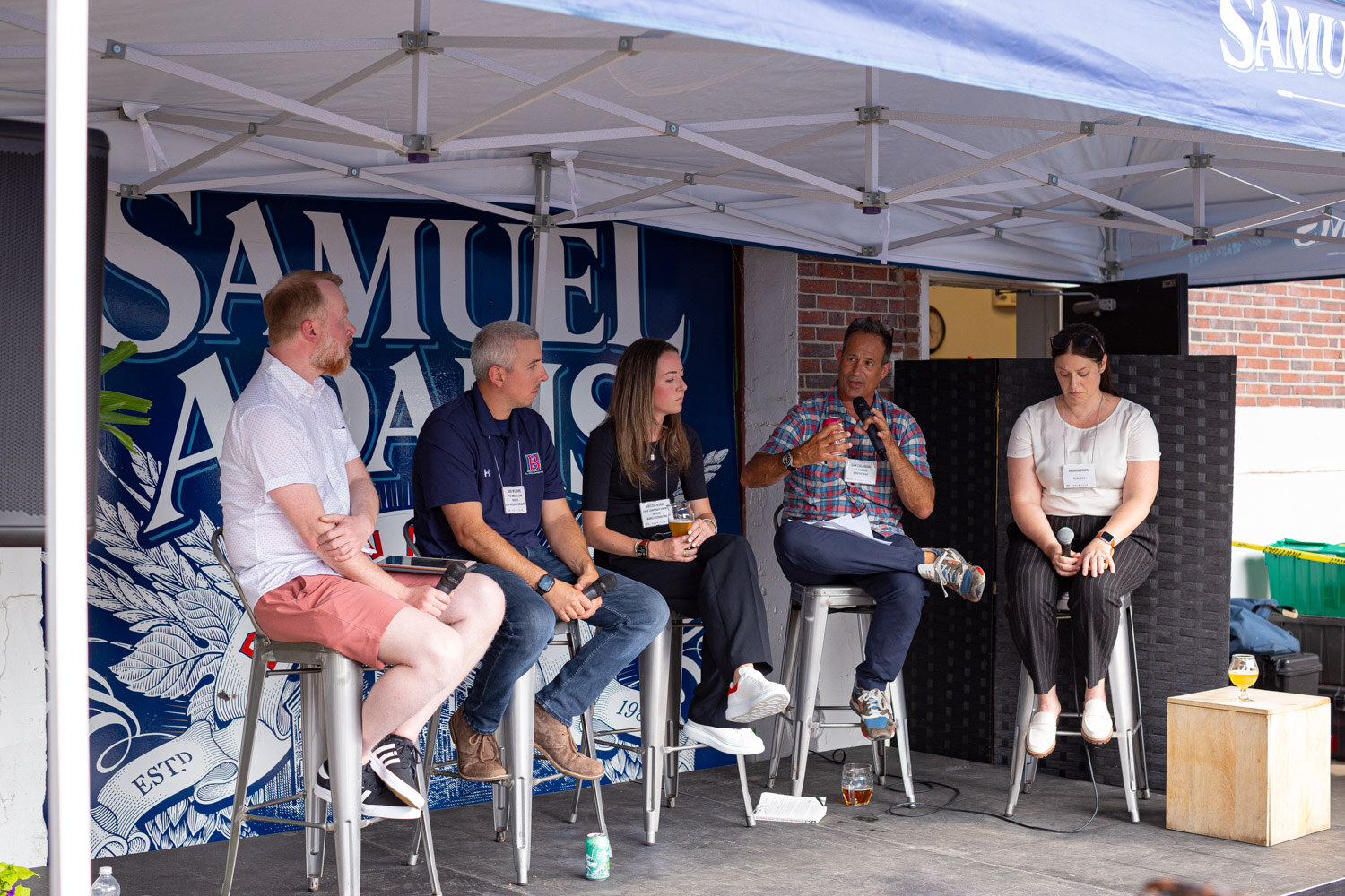 Wide view of five people seated on stools on a stage under a white tent in front of a Samuel Adams mural.