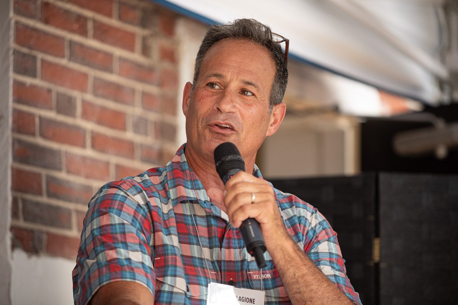 A man in a plaid shirt with a lanyard speaks into a microphone against a brick wall.