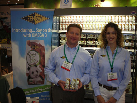A man and a woman in light blue shirts and lanyards stand in front of a ZenSoy banner and a refrigerated display. The man holds small cartons of soy drink.