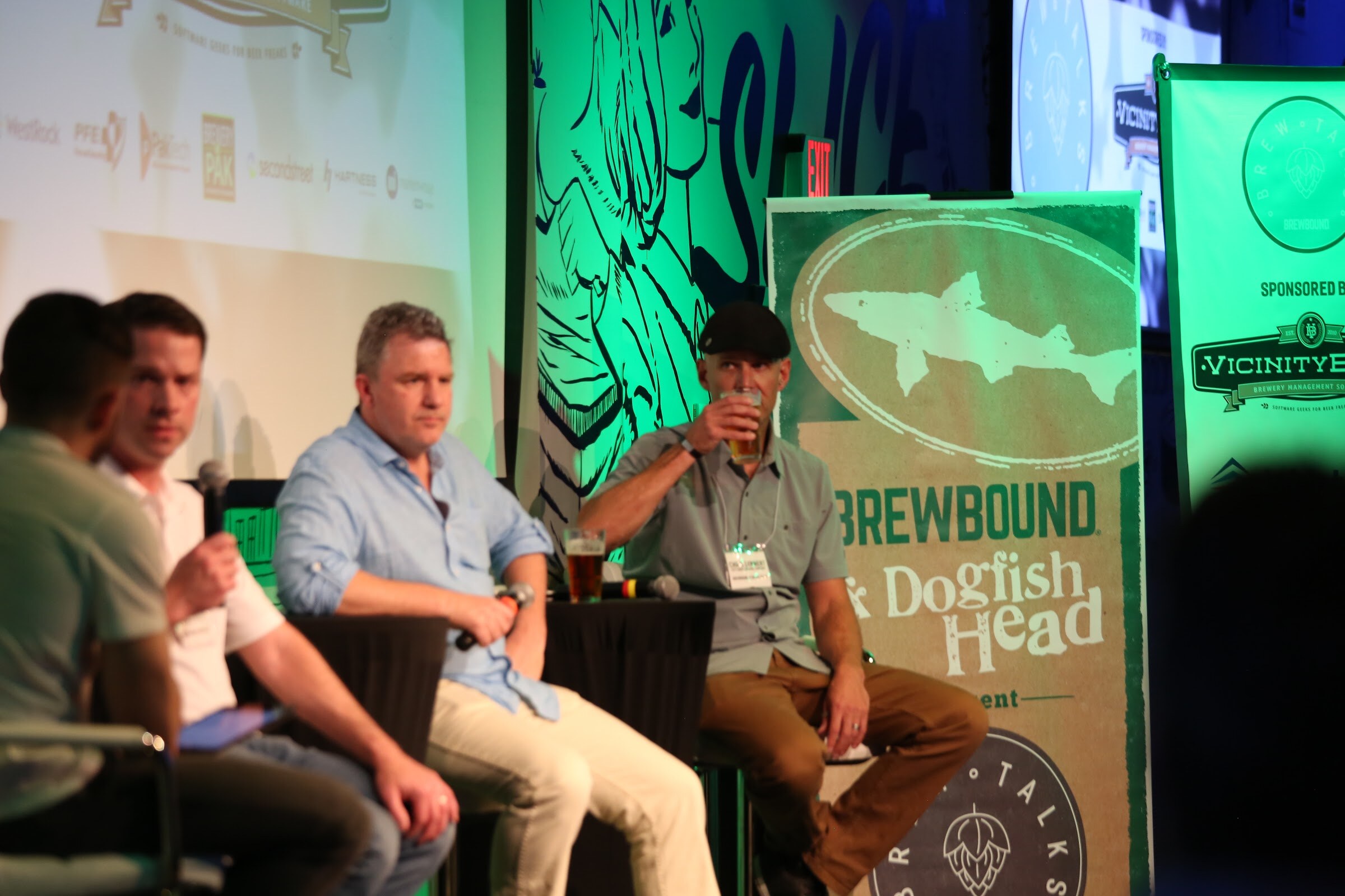 Four people seated on stage stools, one with his back to the camera, beside a green and tan banner reading Brewbound and Dogfish Head.