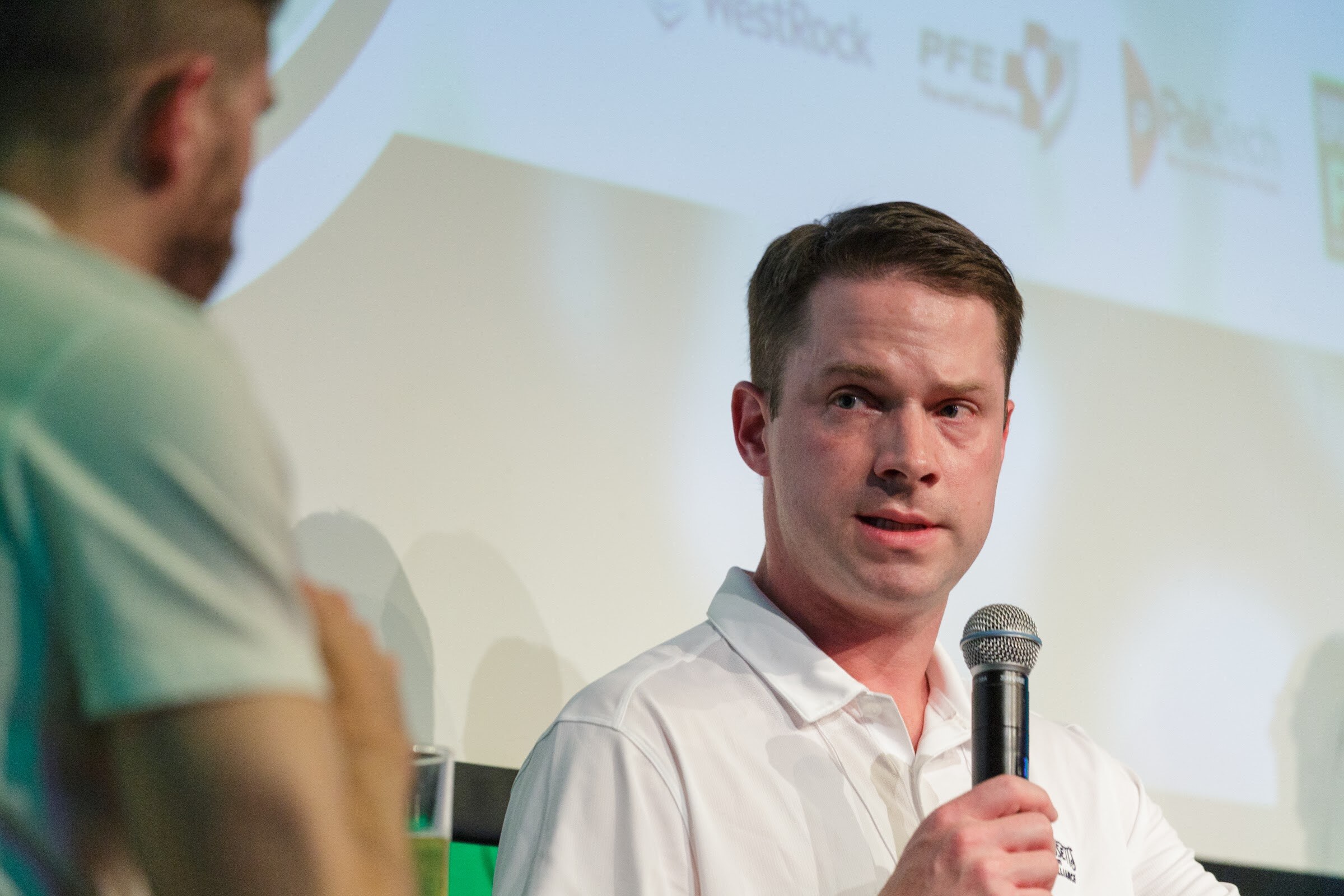 A man in a white polo shirt holds a microphone while another person is partly in frame at left, with a bright projection screen behind.