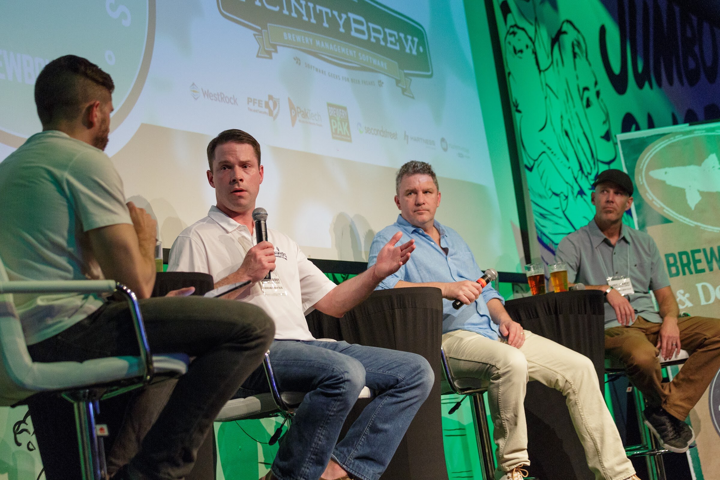 Four men on stage seats facing the audience under a large projection showing the VicinityBrew logo and other sponsor names.