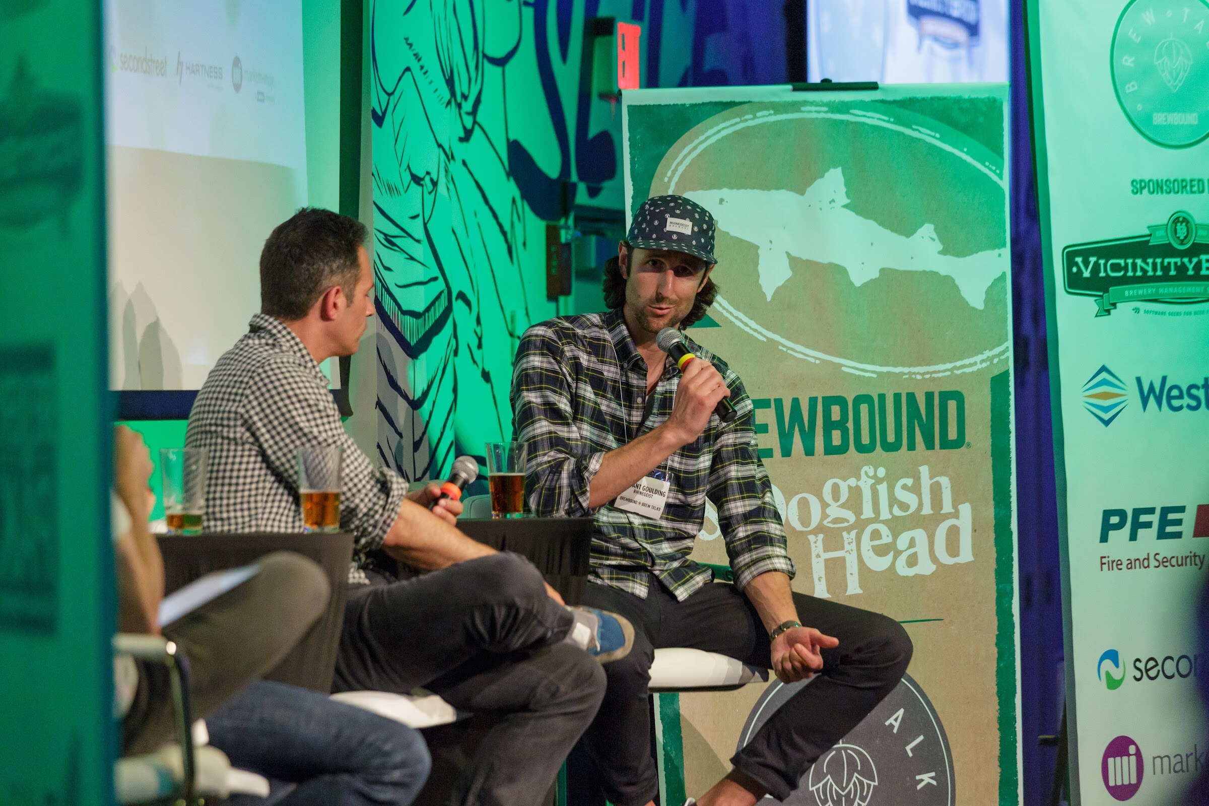 Two men on stage seats, one in a plaid shirt and cap speaking into a microphone, with a Brewbound and Dogfish Head banner behind them.