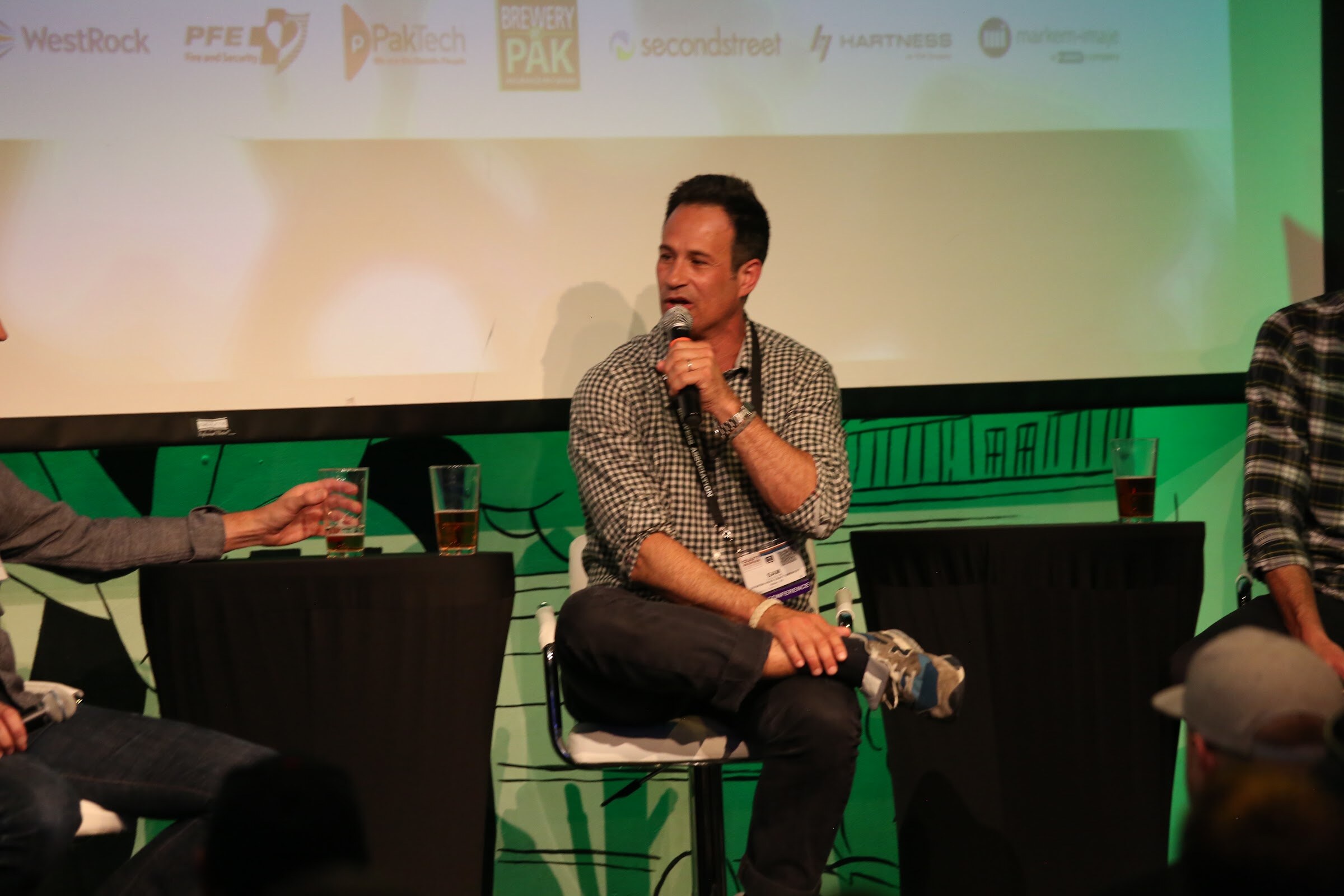A man in a gingham shirt and lanyard holds a microphone, with beer glasses on a draped table and a projection screen behind.