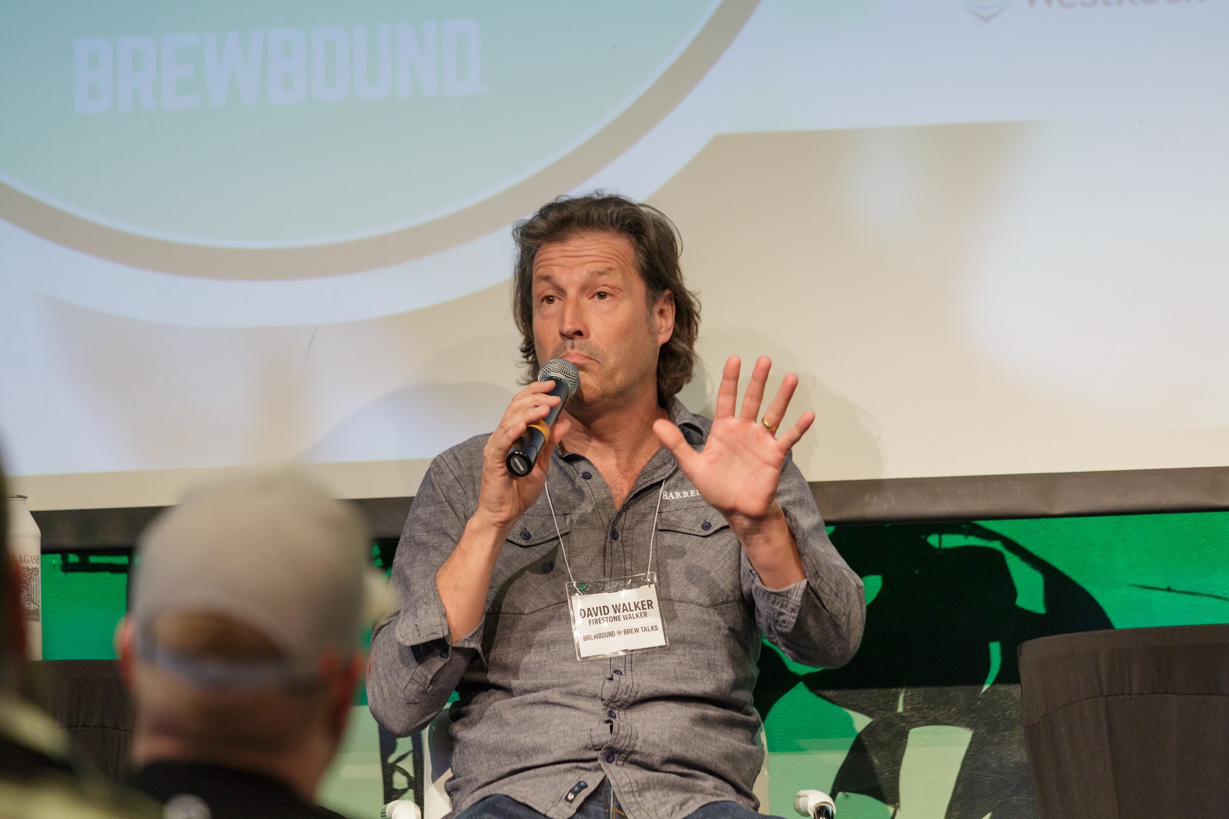 A long-haired man in a gray button shirt and name badge holds a microphone and raises one open hand against a projection screen.