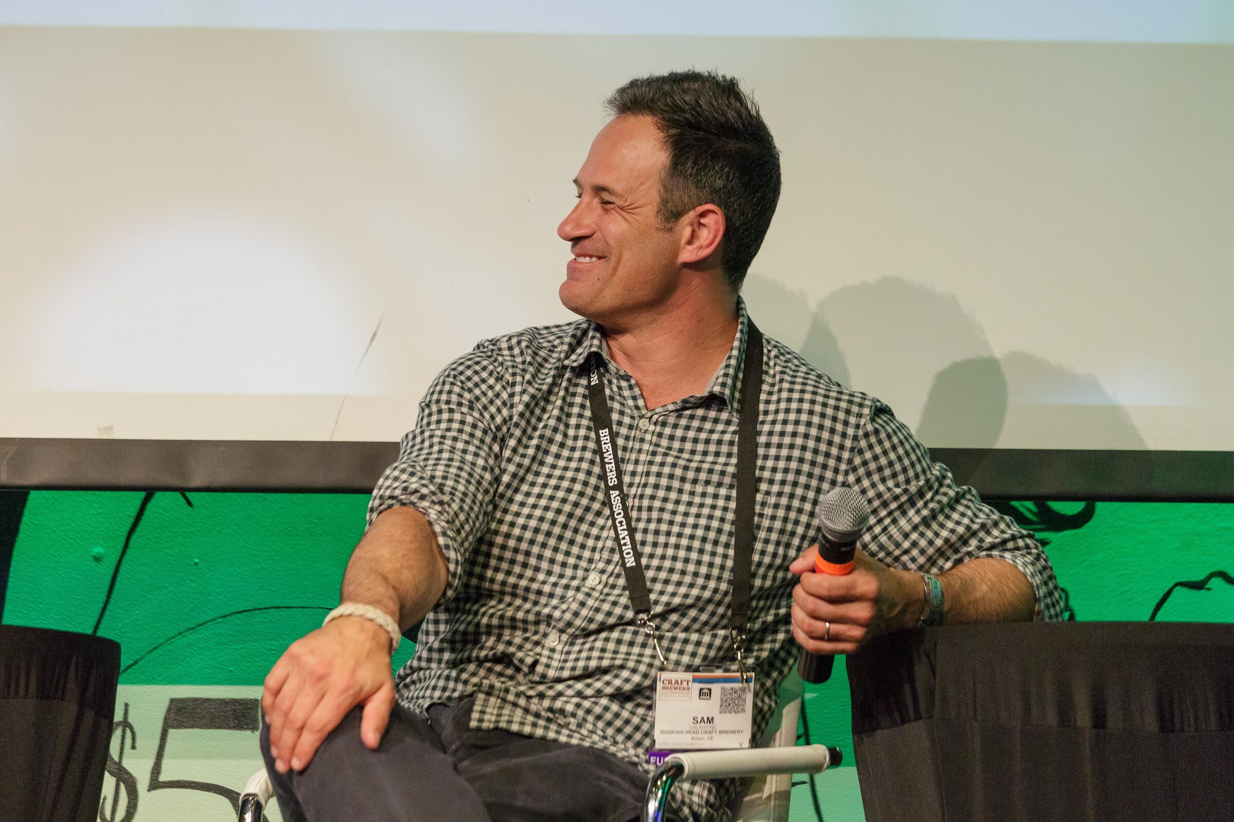 A man in a checked shirt with a conference lanyard sits with a microphone on his knee, turned to the side and smiling.