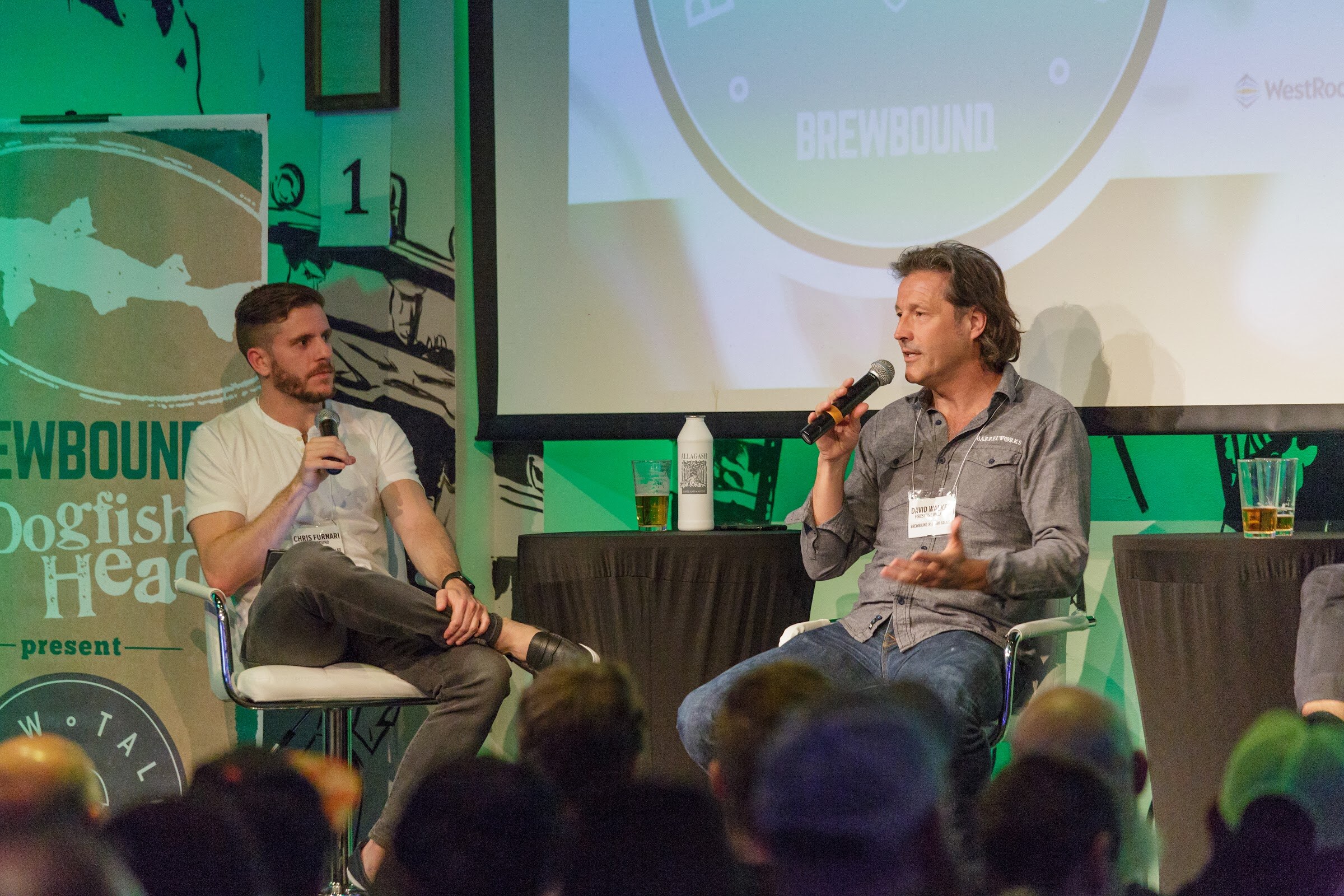 Two men on stage chairs, one in a white shirt holding a microphone and one in a gray shirt speaking, with a Dogfish Head banner at left.