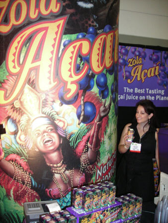 A tall Zola Açaí banner shows a smiling woman in a feathered costume with açaí berries. A woman in black holds a small package beside stacked Zola Açaí boxes and a second banner.