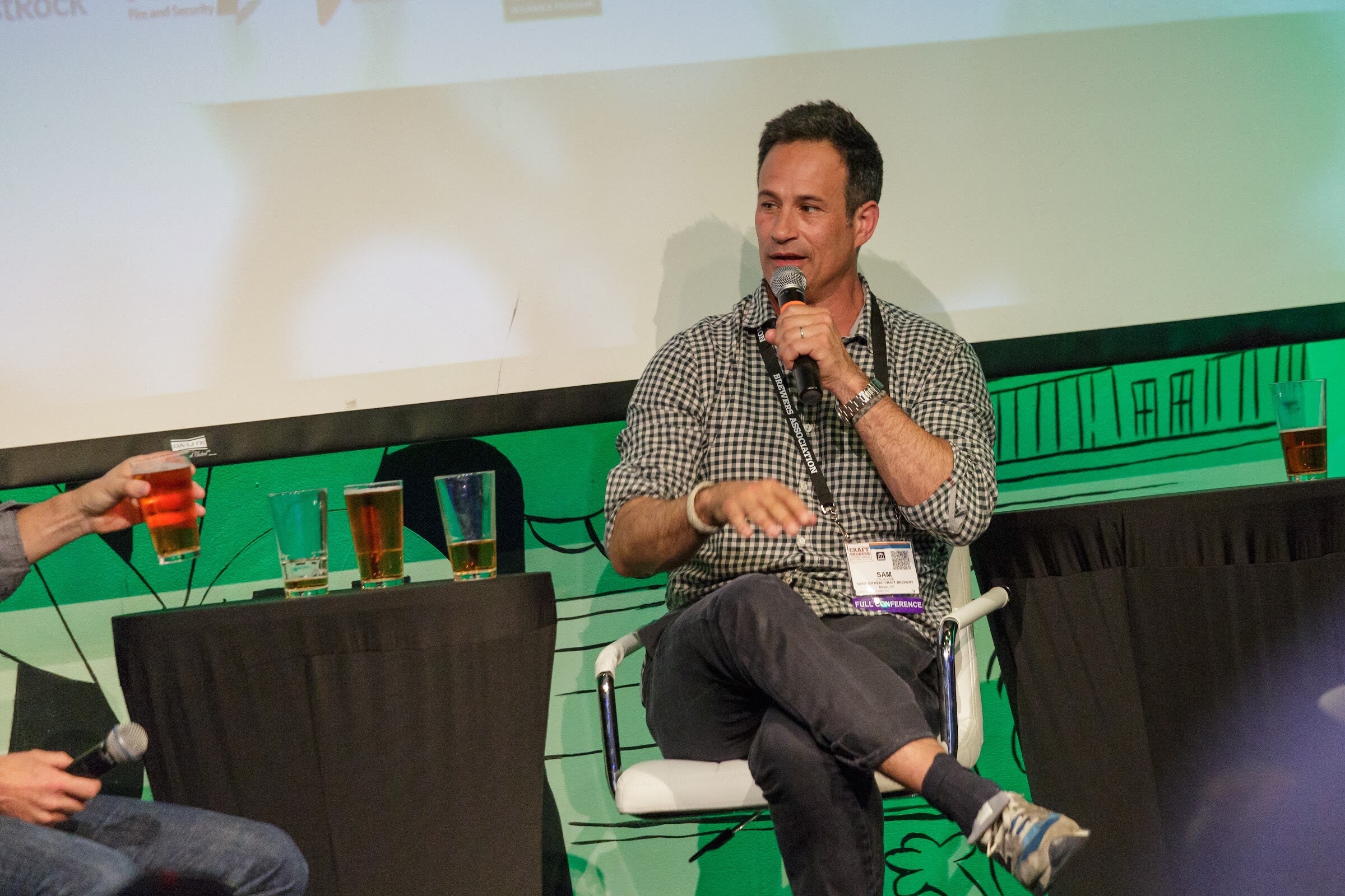 A man in a checked shirt sits cross-legged in a white chair holding a microphone, with glasses of beer on a table covered in black cloth.