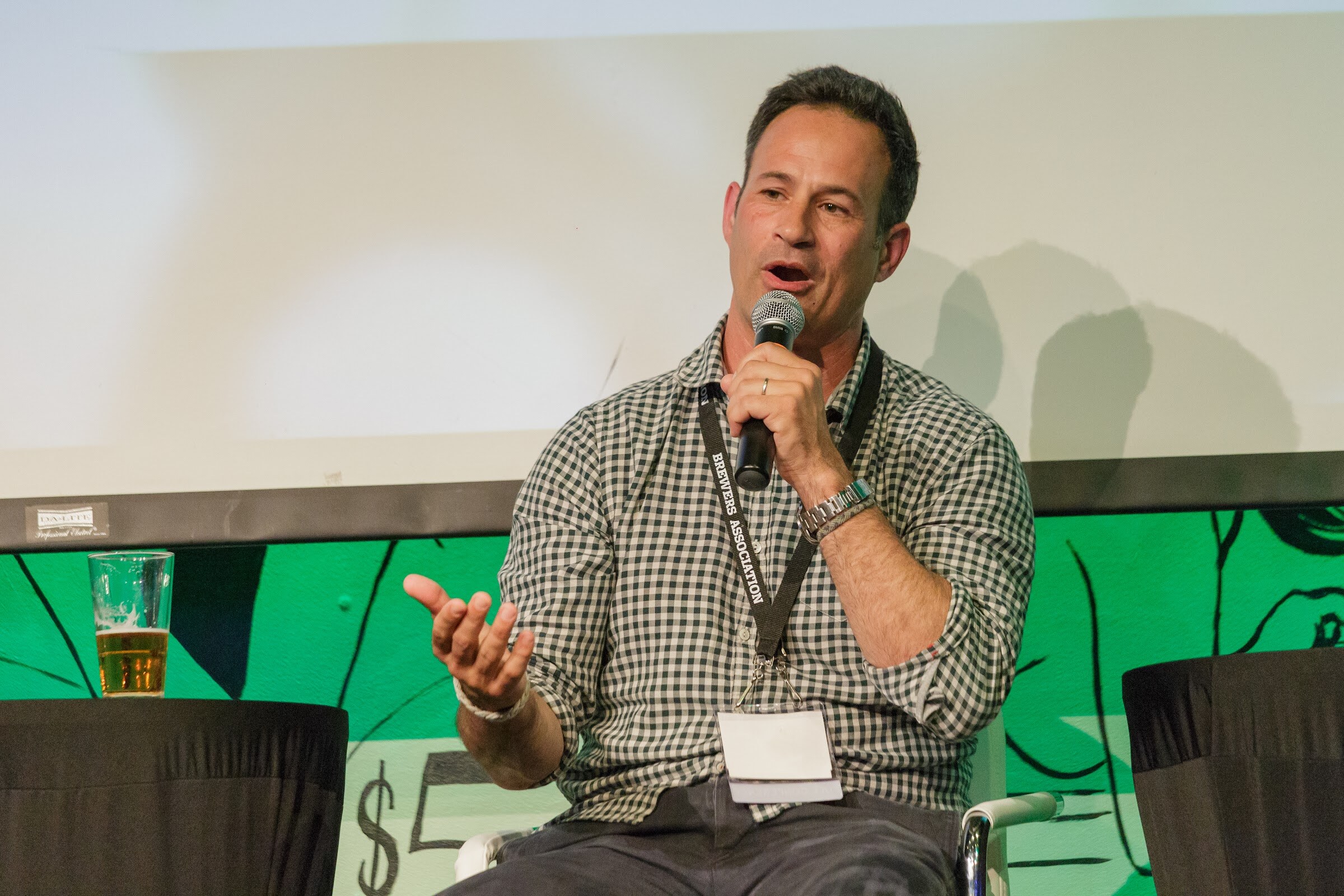 A man in a gingham shirt and lanyard speaks into a microphone with one hand extended, a pint glass on the table beside him.
