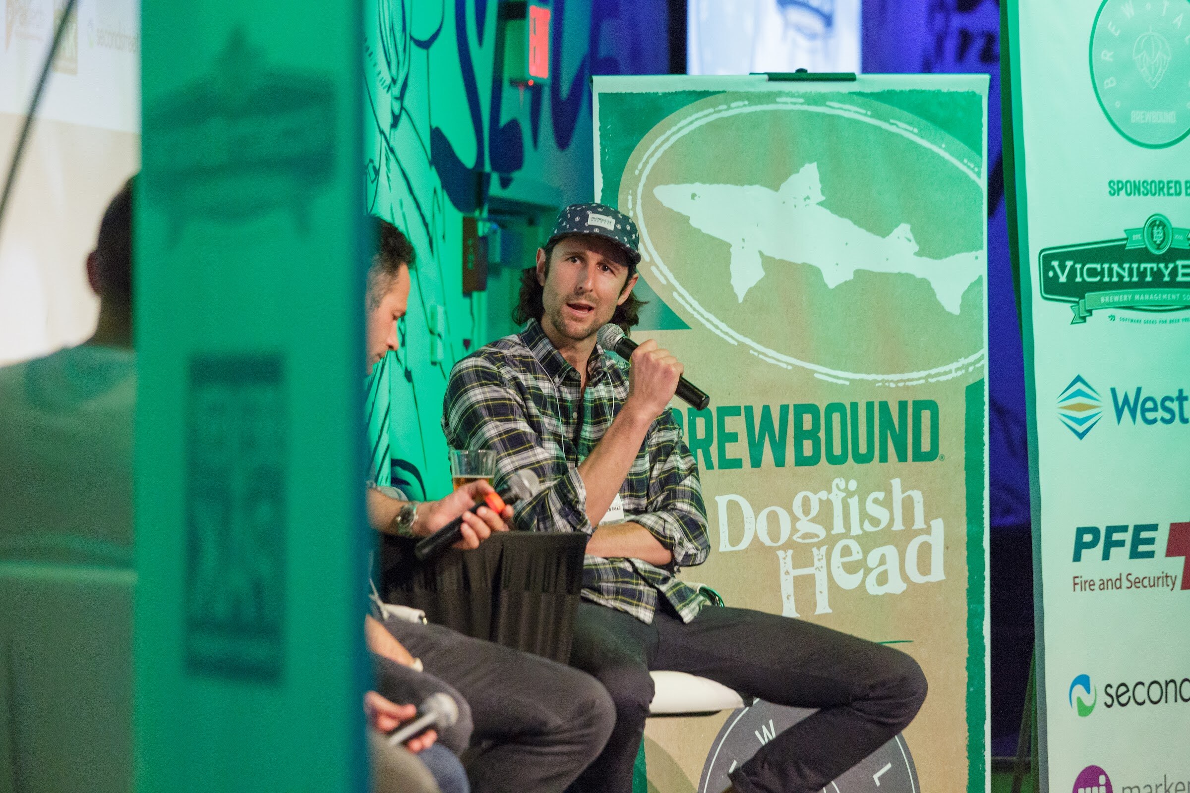 A man in a plaid shirt and patterned cap speaks into a microphone, framed by a Brewbound and Dogfish Head banner and a teal panel in the foreground.