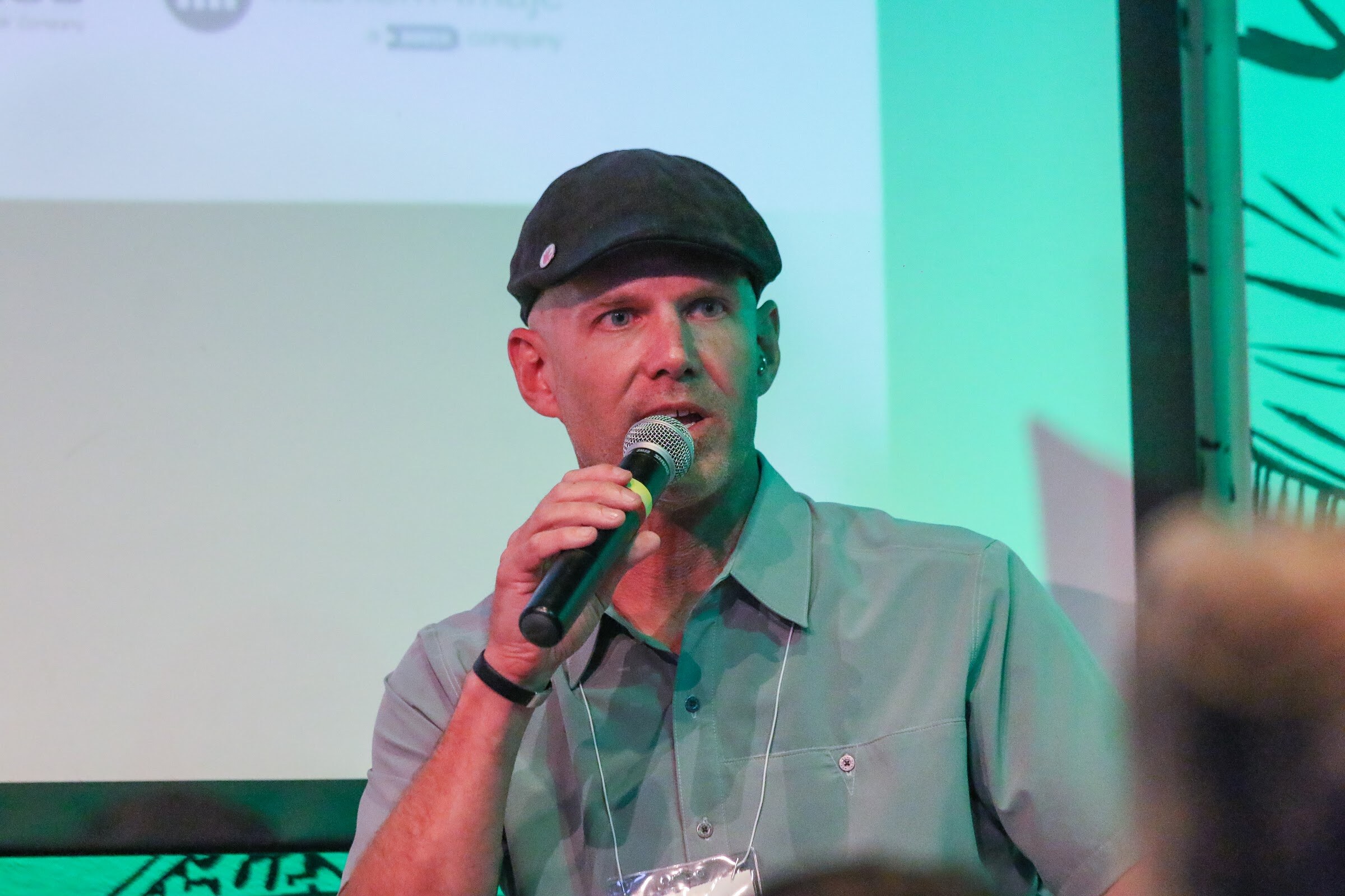 A man in a flat cap and gray collared shirt with a lanyard speaks into a microphone in front of a green and white backdrop.